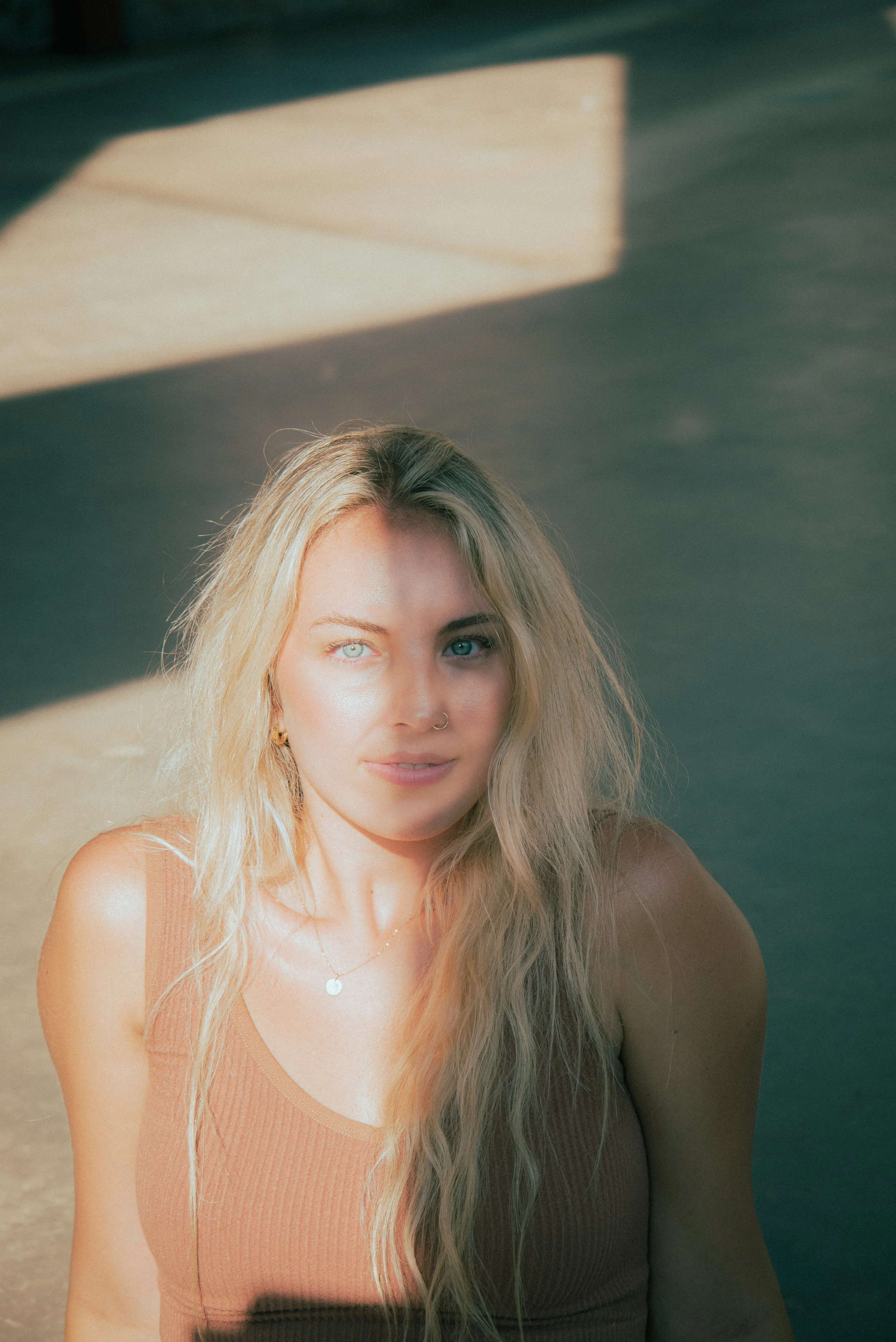 A young woman with blonde hair and blue eyes sitting outside, wearing a peach-colored tank top and a silver necklace, with sunlight and shadow patterns on her face.
