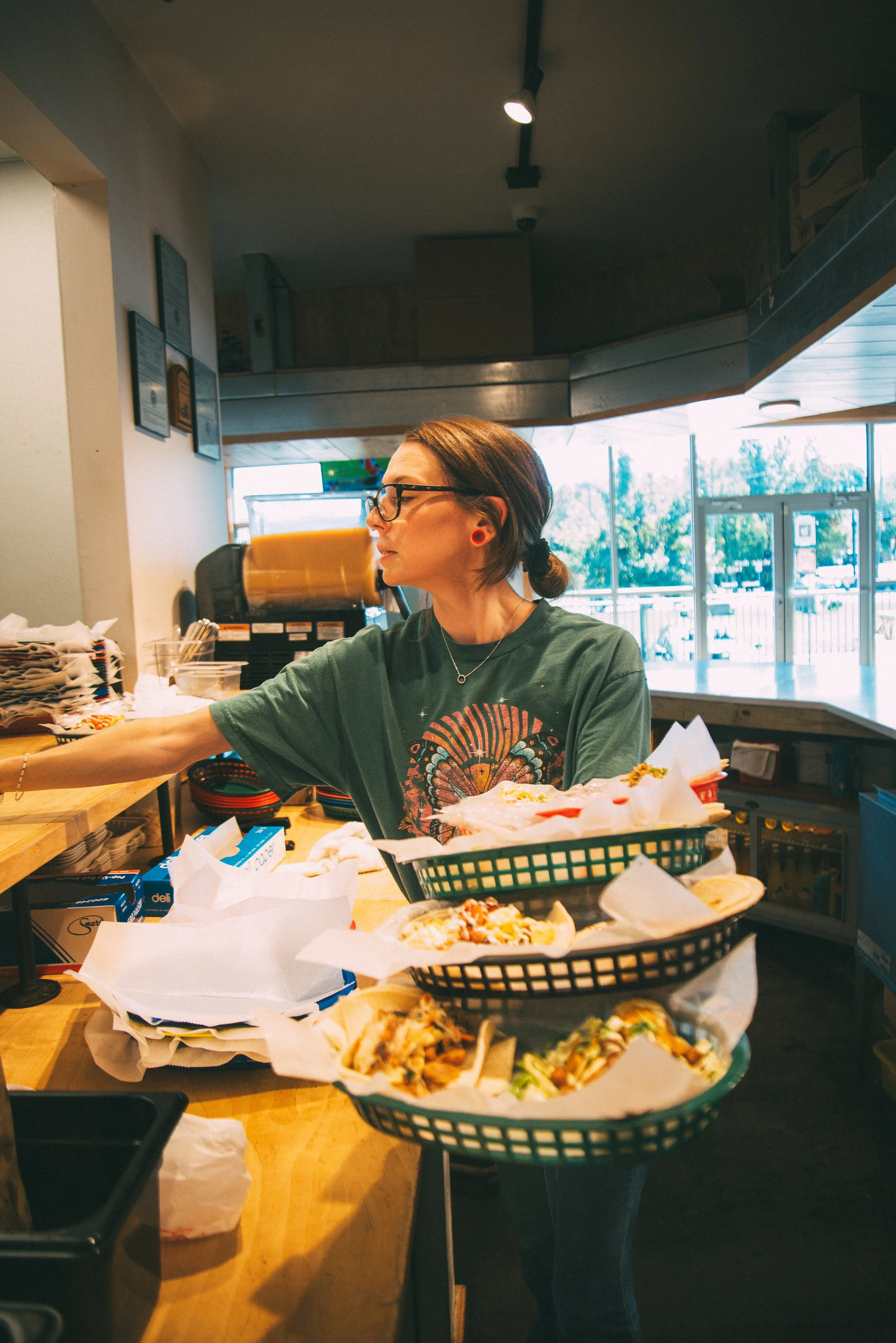 A woman with glasses and earrings serving herself food at a restaurant counter, with trays of pizza, and a window showing trees outside.
