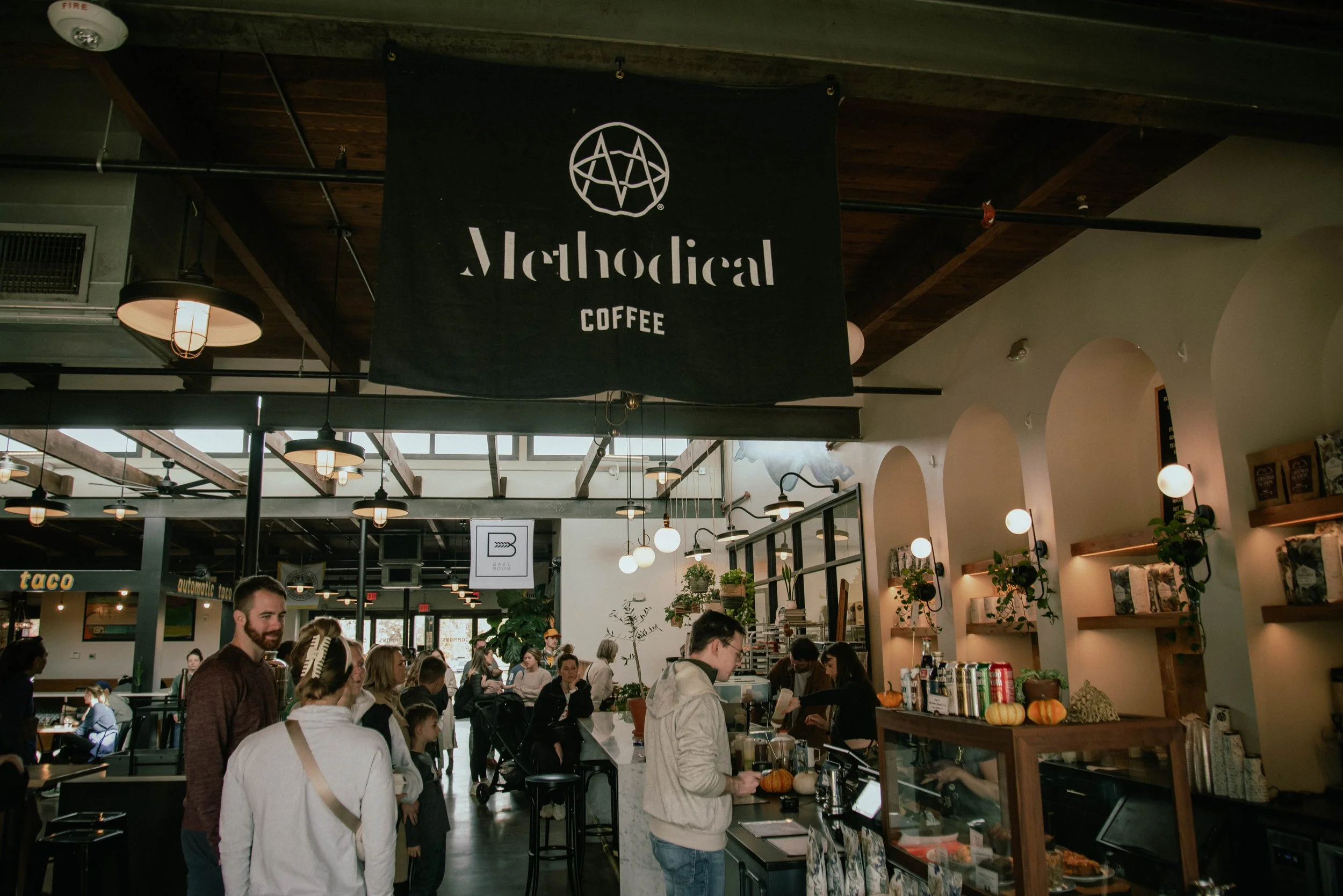 Interior of a busy coffee shop with a black sign reading 'Metropolitan Coffee' hanging from the ceiling, people ordering and standing in line, warm lighting, and decorative plants.