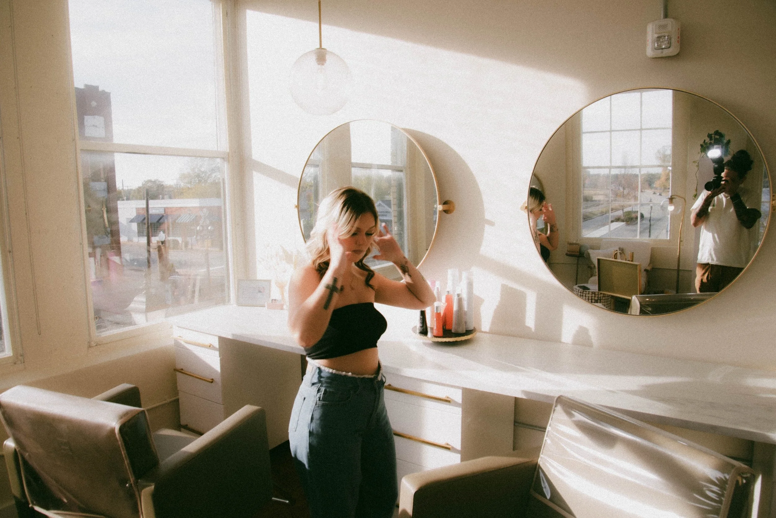 A woman with long blonde hair, wearing a black tube top and high-waisted jeans, stands in a brightly lit room with large windows. She is adjusting her hair. Behind her, a photographer with a camera and flash is taking her picture, and her reflection 