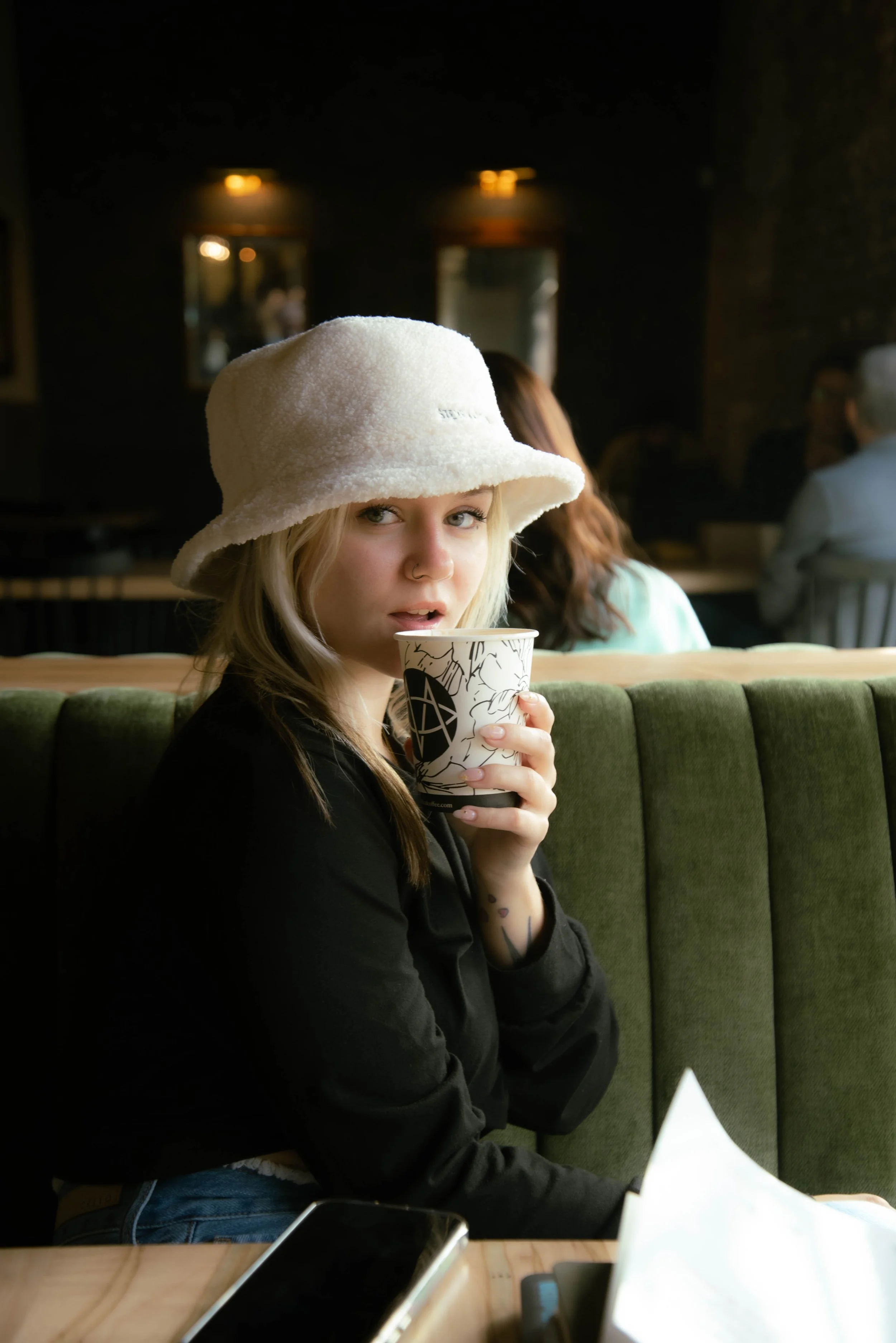 A young woman with blonde hair, blue eyes, and a nose ring holding a black and white patterned cup, sitting in a coffee shop wearing a white fuzzy hat and a black jacket.