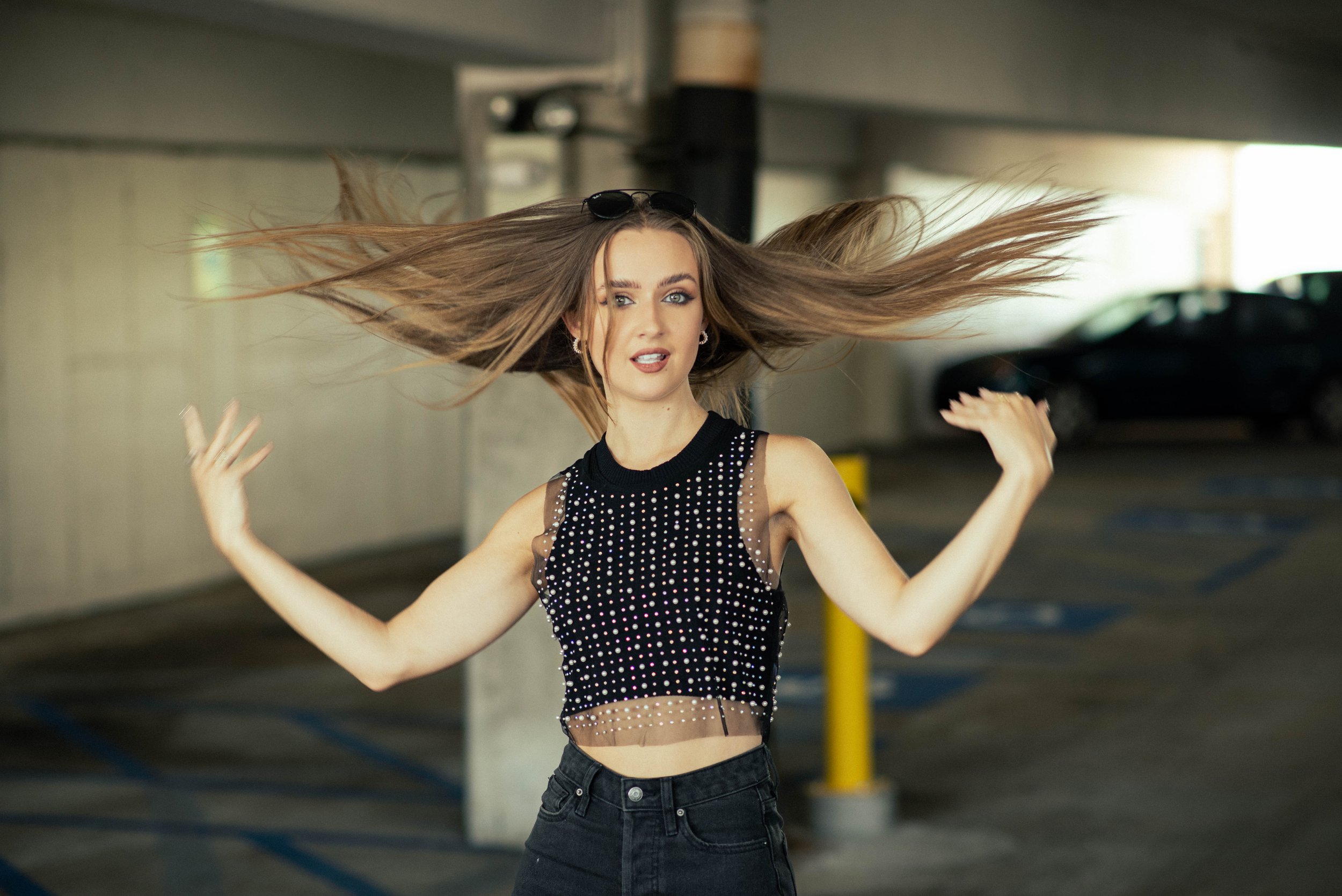 A young woman with long flowing hair, wearing sunglasses on her head, a sleeveless black top with white polka dots, and black jeans, is dancing or posing in an indoor parking garage.