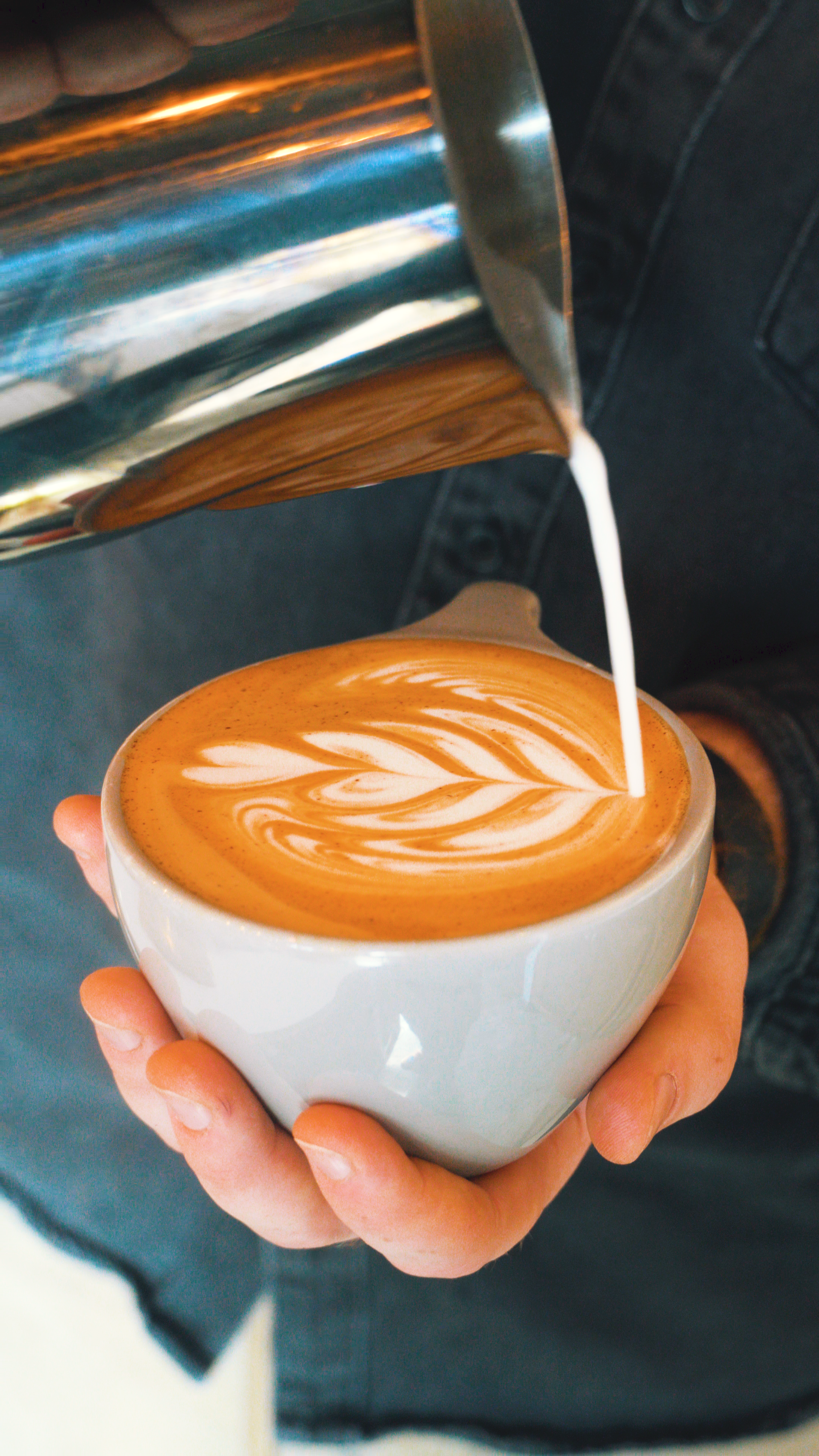 Person holding a white cup of latte with a leaf pattern in the foam, while pouring milk from a metal pitcher.