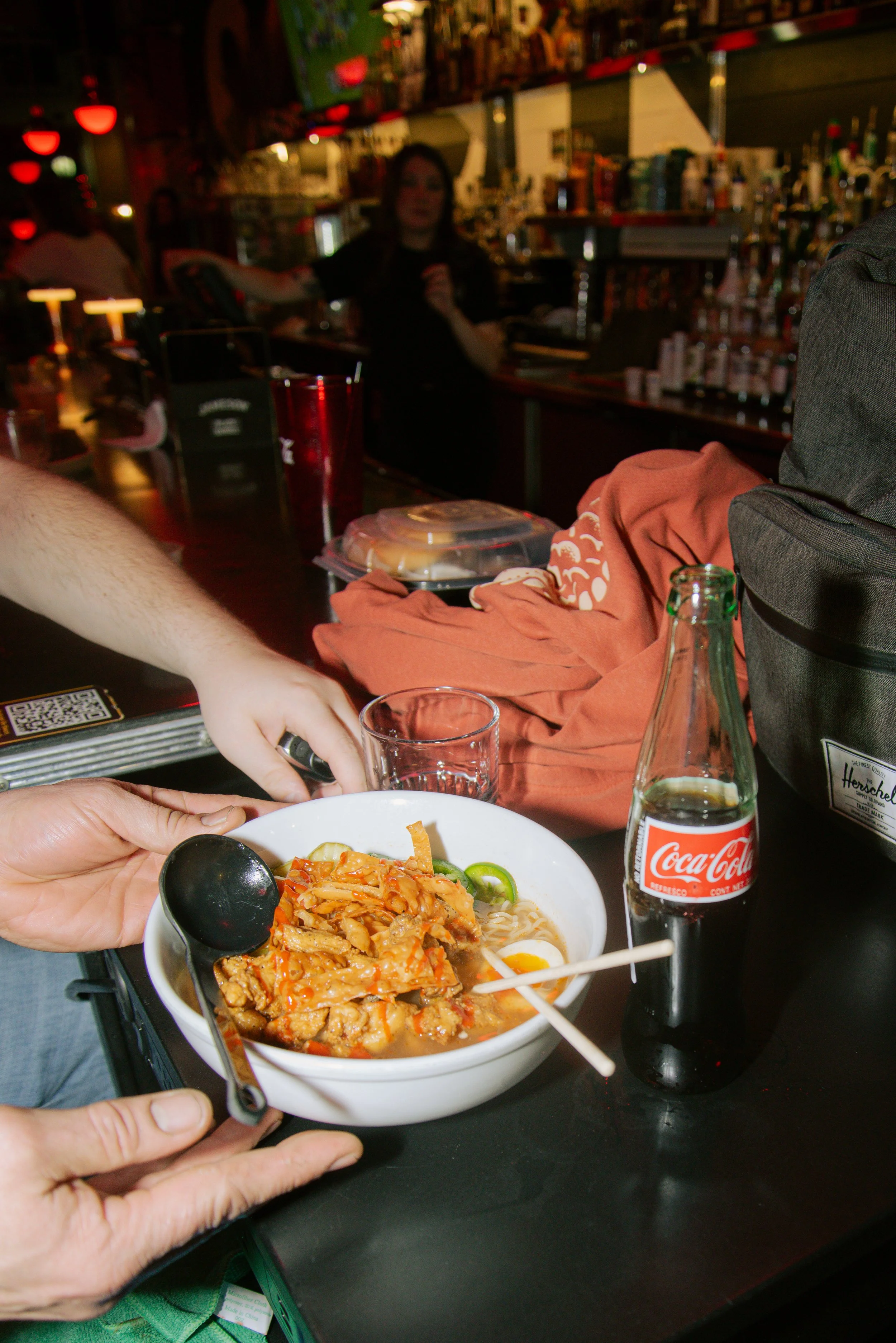 A bowl of Asian-style noodle soup with vegetables and meat, alongside a glass bottle of Coca-Cola, on a black table at a restaurant.