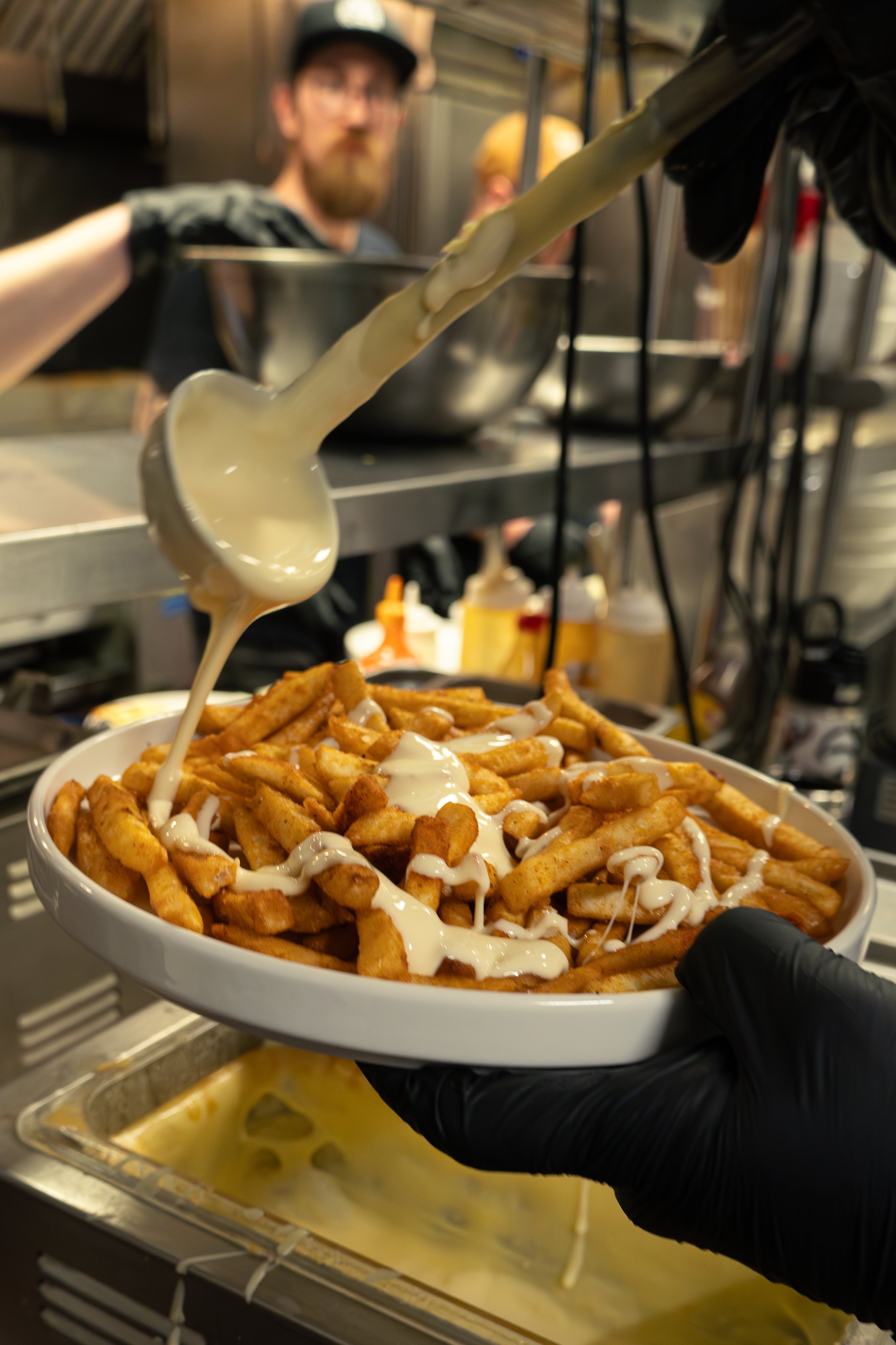 A person with black gloves is holding a bowl of French fries topped with cheese sauce and drizzled with white sauce. In the background, someone is preparing more cheese sauce in a metal container, with a man with a beard and glasses observing.
