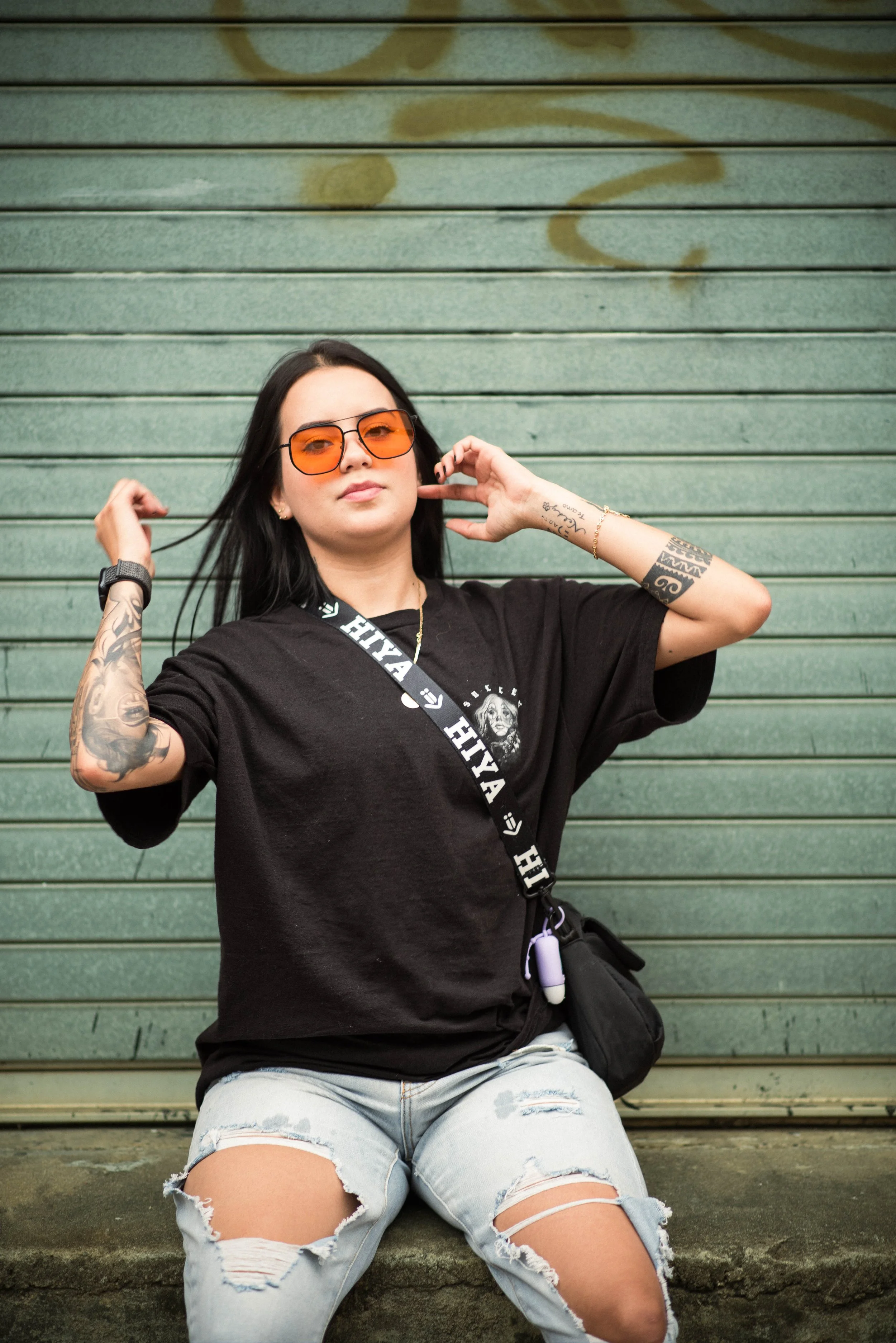 A woman with black hair wearing orange sunglasses, a black t-shirt with a graphic, and light ripped jeans, sitting against a green metal shutter with graffiti, adjusting her hair with both hands.
