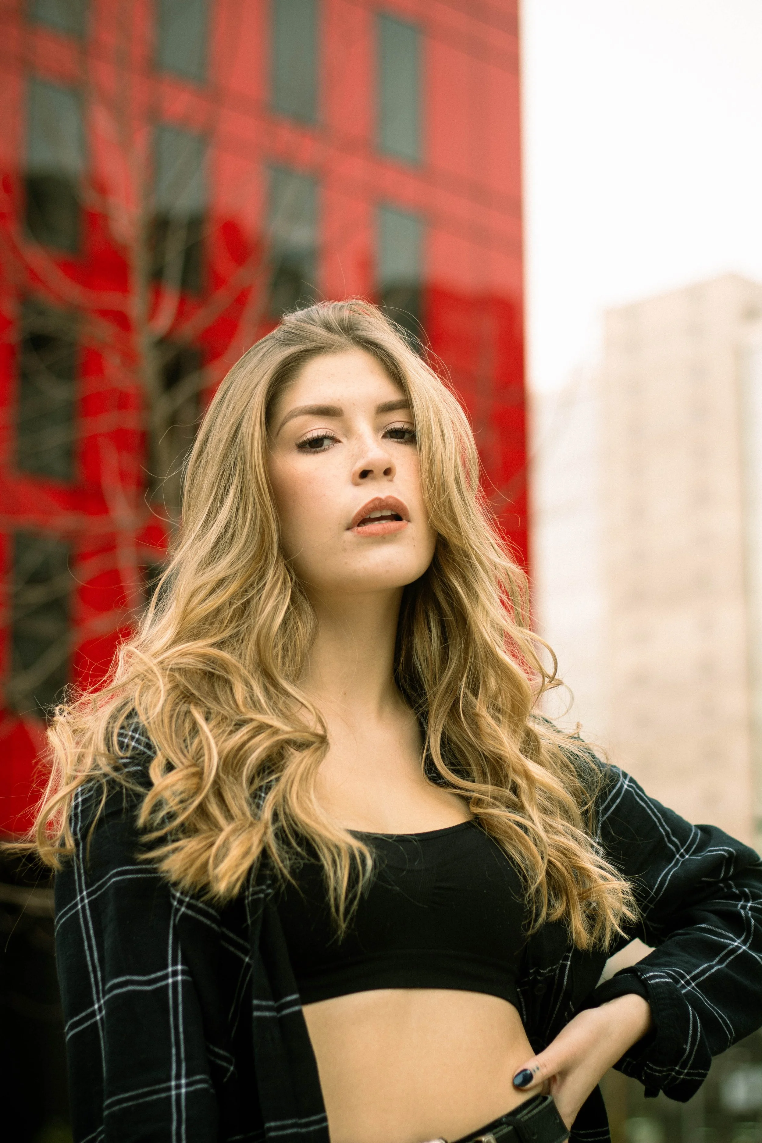 A young woman with long, wavy blonde hair standing outdoors in an urban setting. She is wearing a black crop top and a black plaid shirt, with one hand on her waist. The background features a red brick building and some trees.