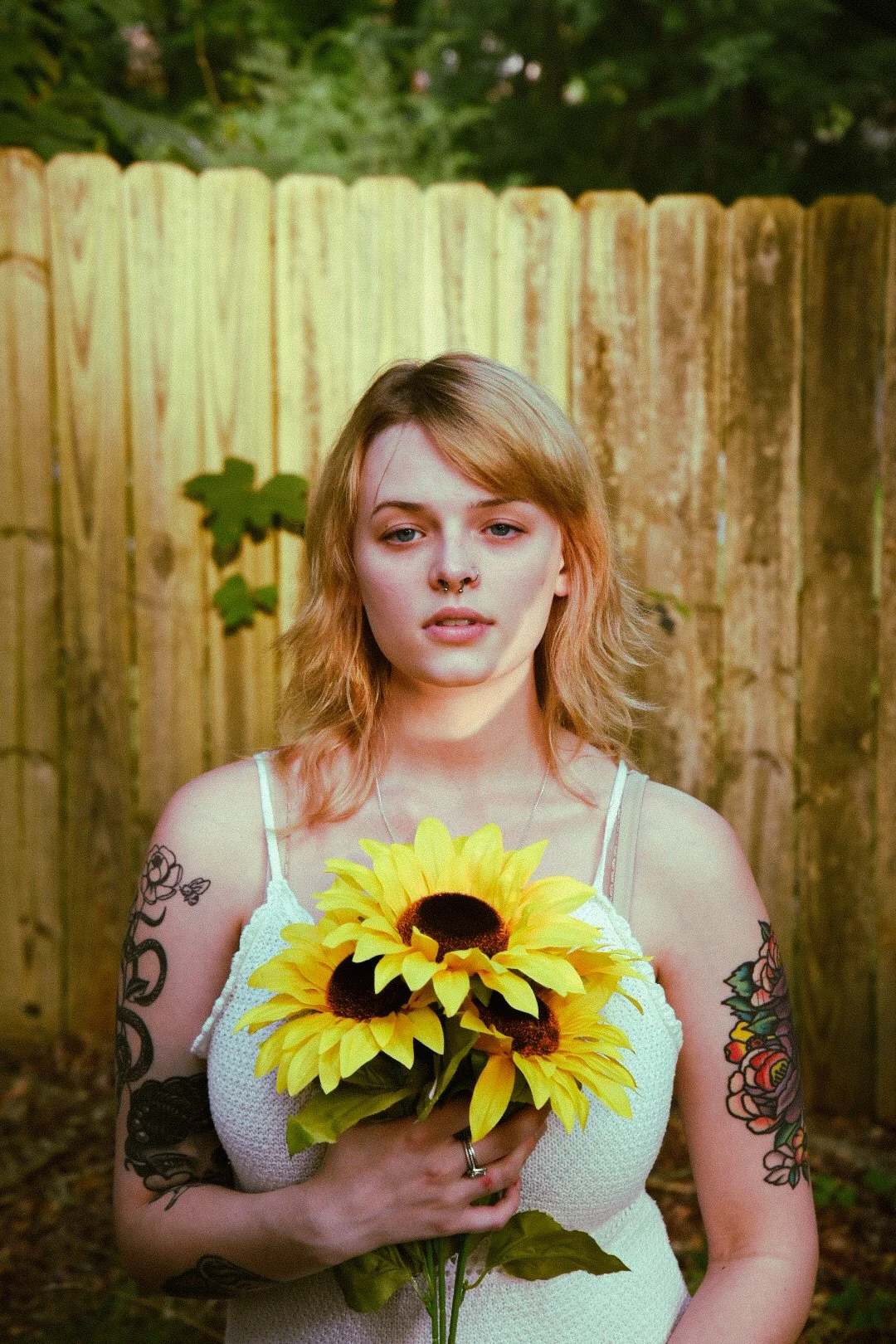 A young woman with short, wavy, blonde hair and tattoos on her arms stands outdoors in front of a wooden fence, holding a bouquet of bright yellow sunflowers and looking into the camera.