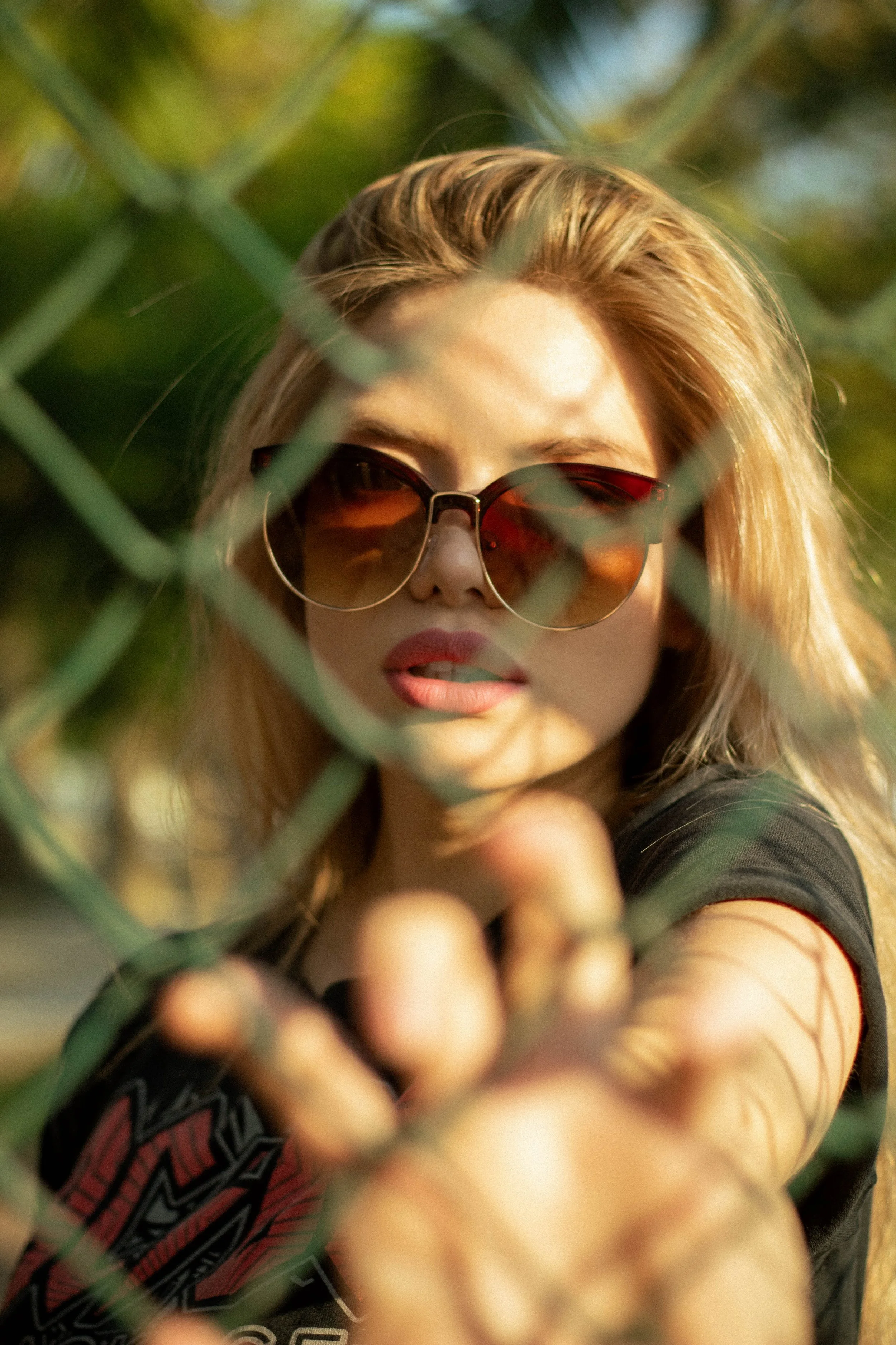 A woman with blonde hair and sunglasses peering through a chain-link fence.