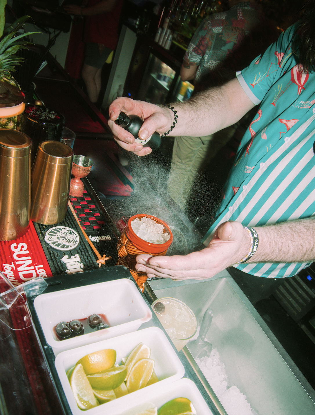 A bartender in a colorful Hawaiian shirt sprinkles crushed ice into a drink at a bar. There are lemon and lime wedges, black cherries, and various bar tools and bottles visible on the counter.