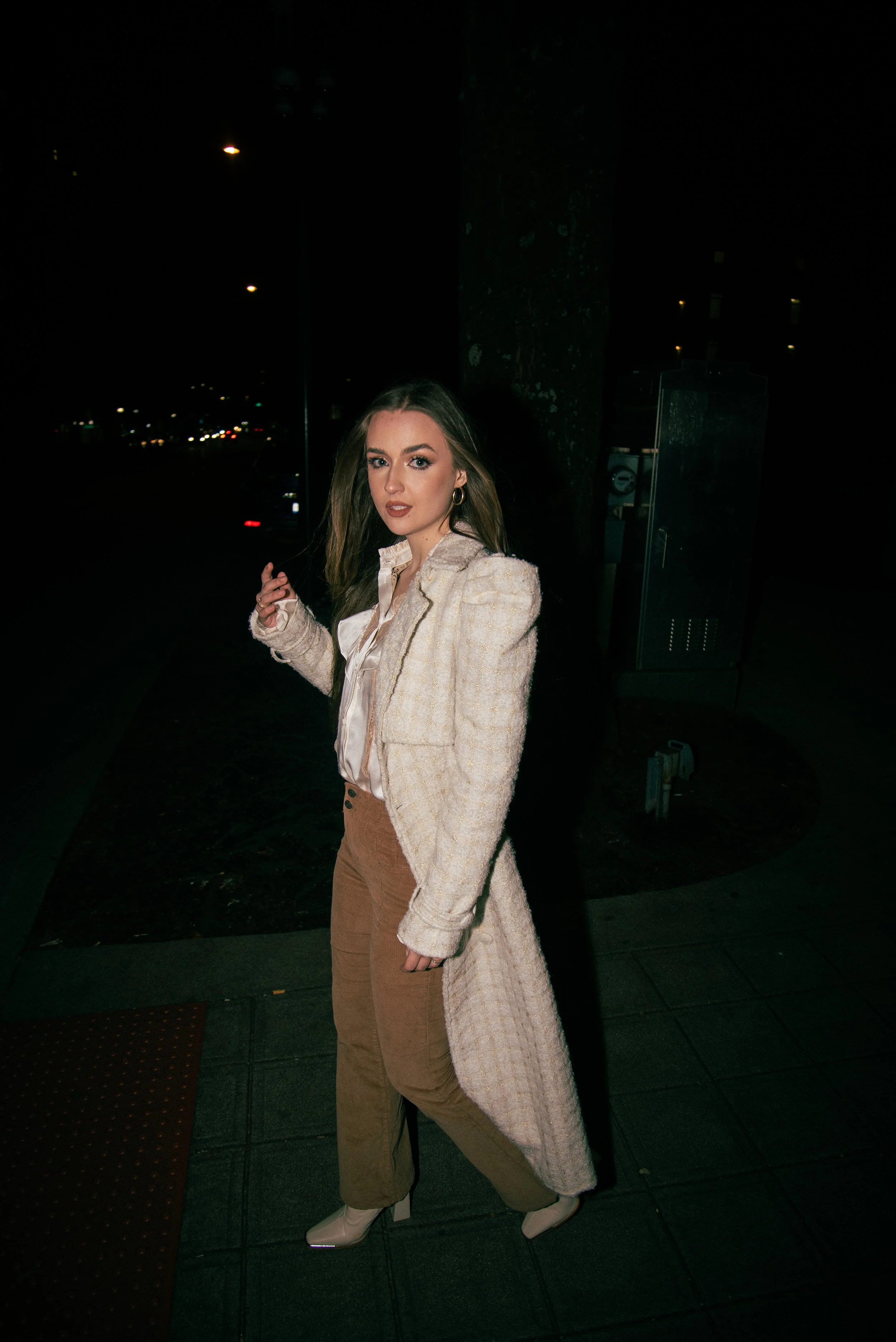 Young woman with long hair, wearing a beige coat, brown pants, and white boots, standing outdoors at night with city lights in the background.