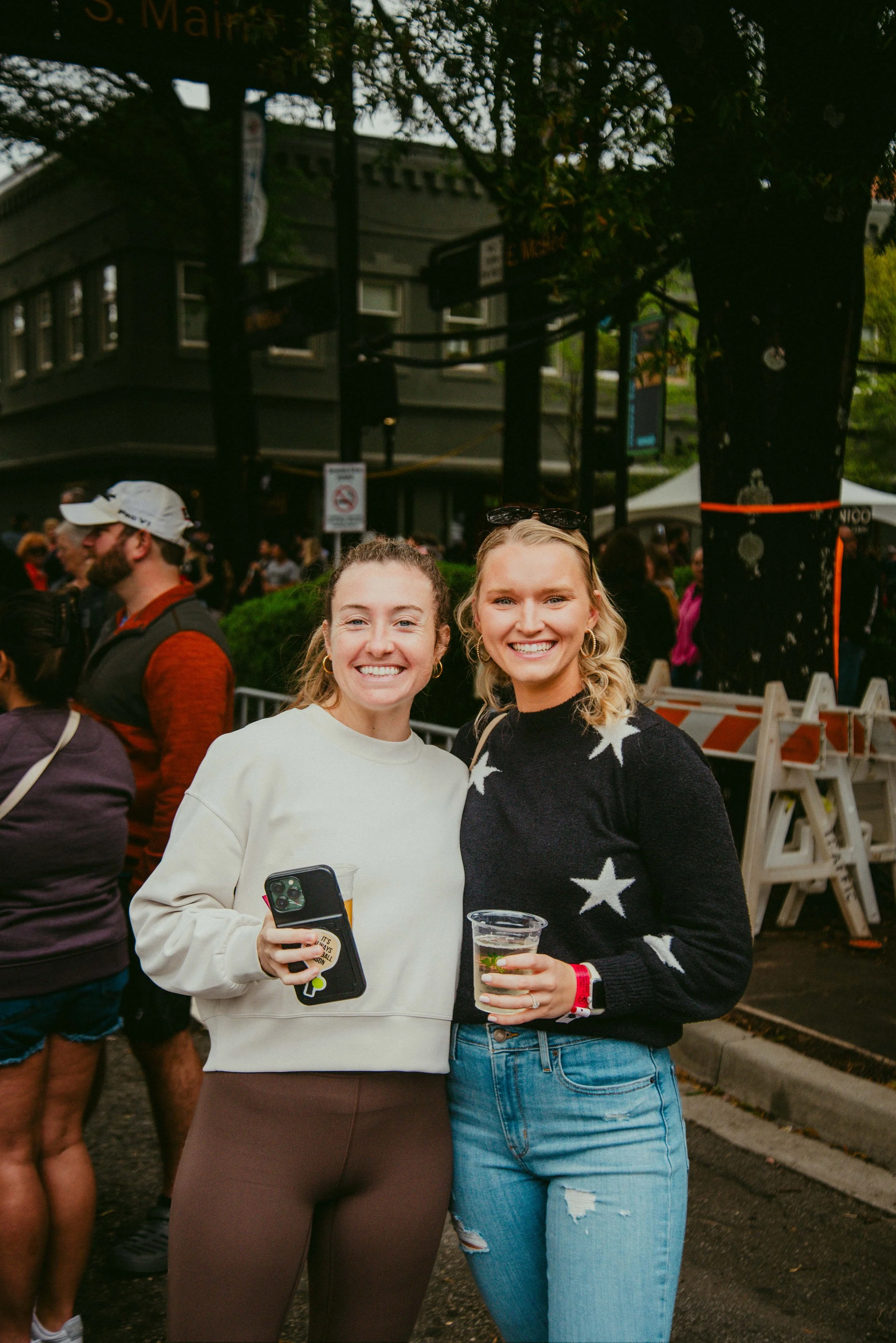 Two smiling women standing together at an outdoor event, holding drinks in clear cups. The woman on the left has light brown hair, is wearing a white sweatshirt and brown pants, and is holding a phone. The woman on the right has blonde hair, is weari