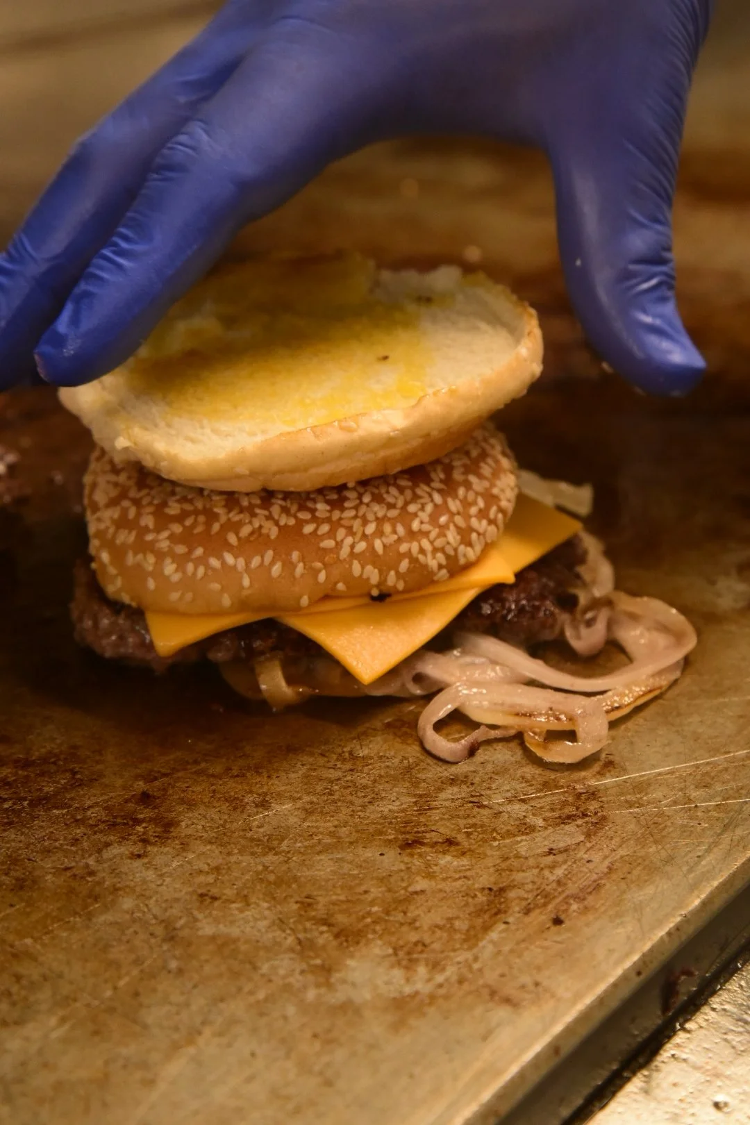 A person wearing a blue glove assembling a sandwich with sesame seed bun, cheese, grilled hamburger patty, onions, and a sliced sandwich bread on top.