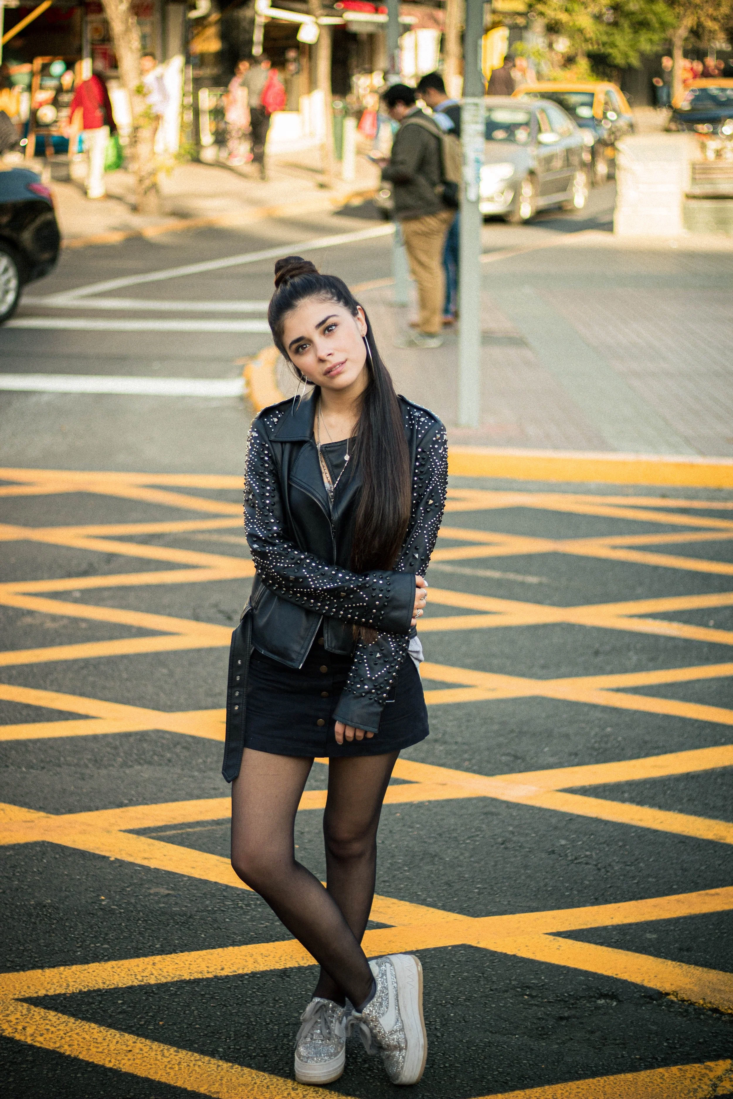 Young woman with long dark hair, wearing a black leather jacket with metal studs, black skirt, sheer black tights, and glittery sneakers, stands on a crosswalk in an urban street during daytime, with cars and pedestrians in the background.