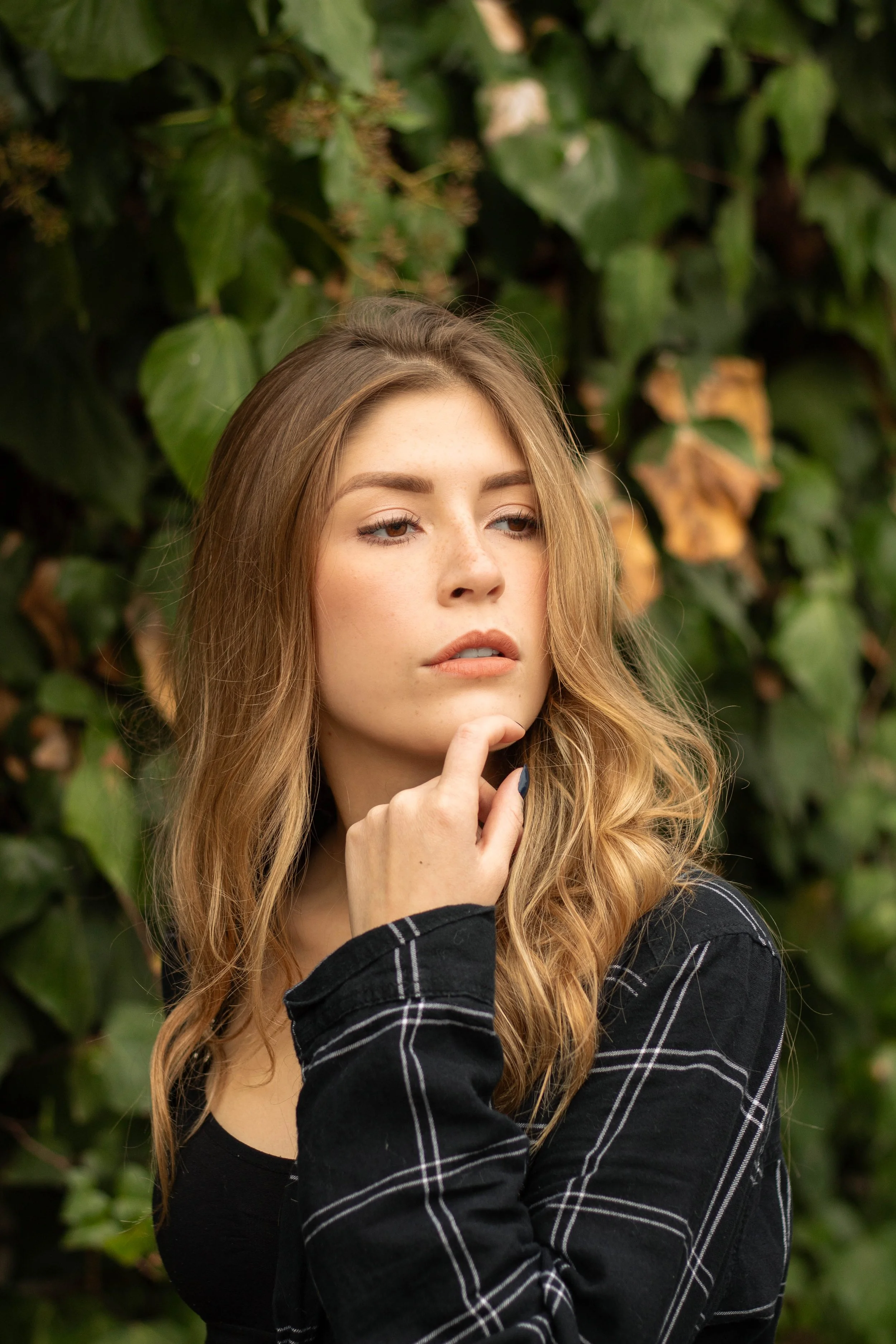 A young woman with long, wavy, light brown hair, wearing a black shirt with white checkered pattern, standing in front of green leafy background, touching her chin with her right hand.