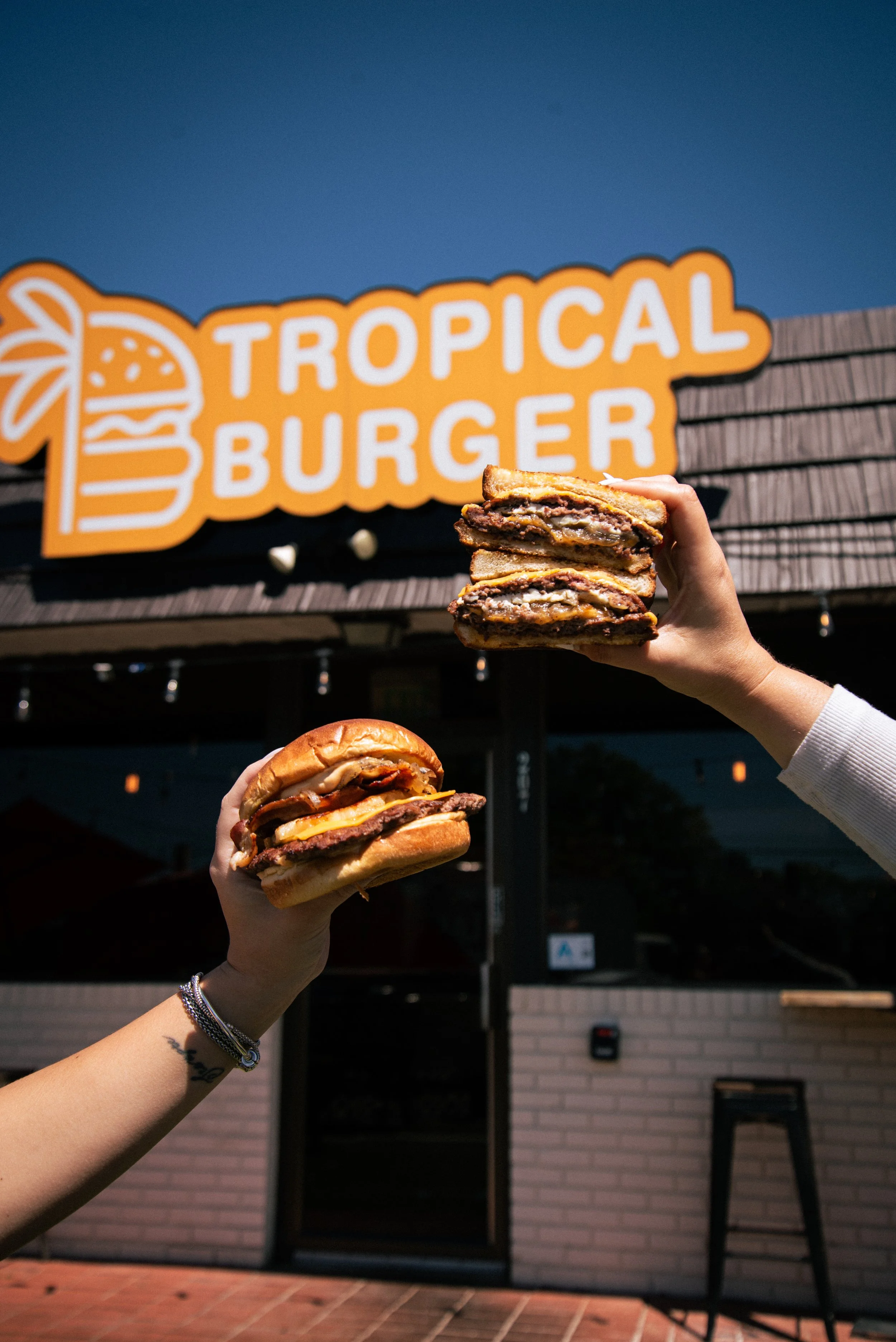 Two hands holding double-stacked burgers in front of a tropical burger sign outside a restaurant.
