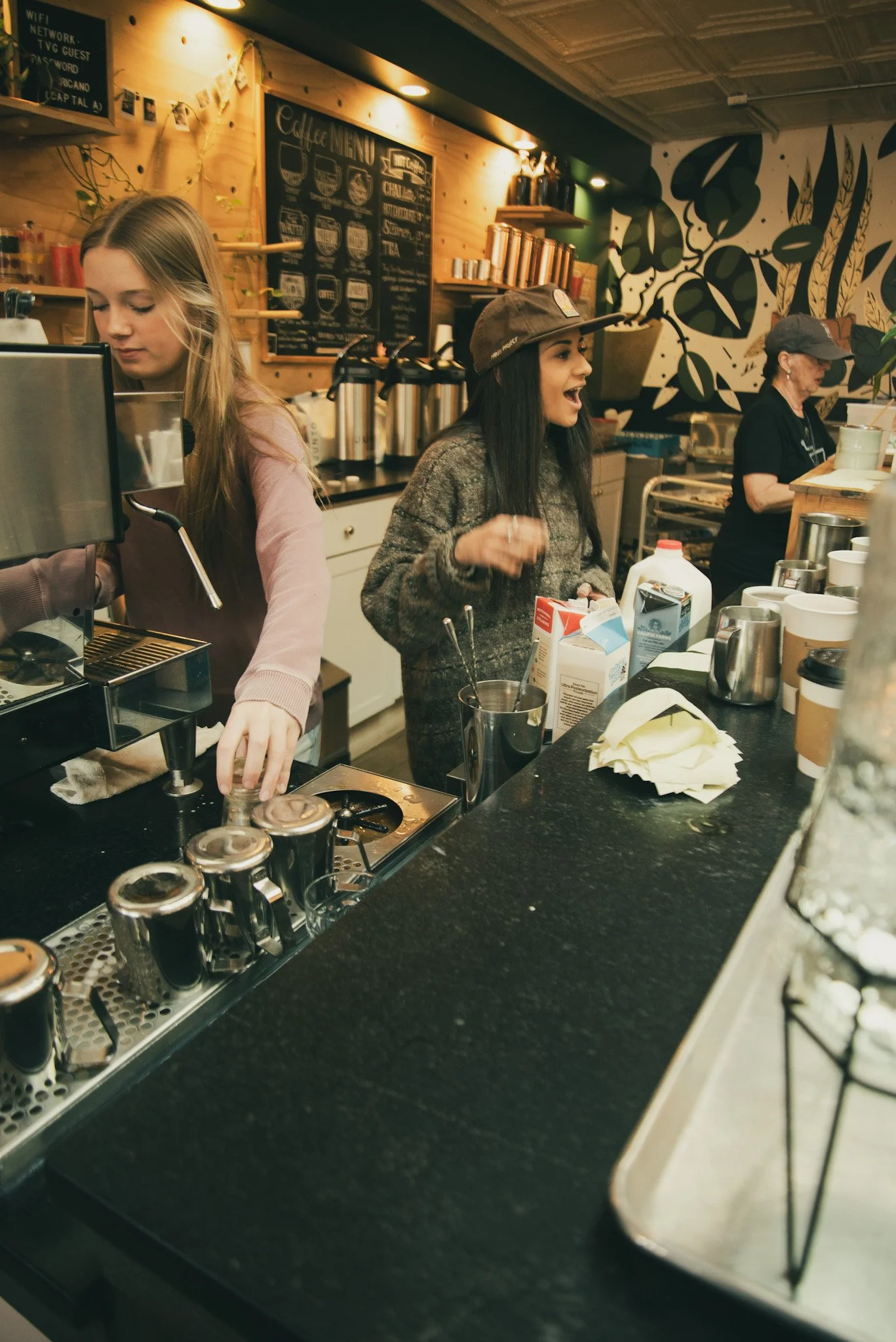 A coffee shop counter with baristas preparing drinks, a menu board on the wall, and various coffee-making equipment and supplies on the counter.