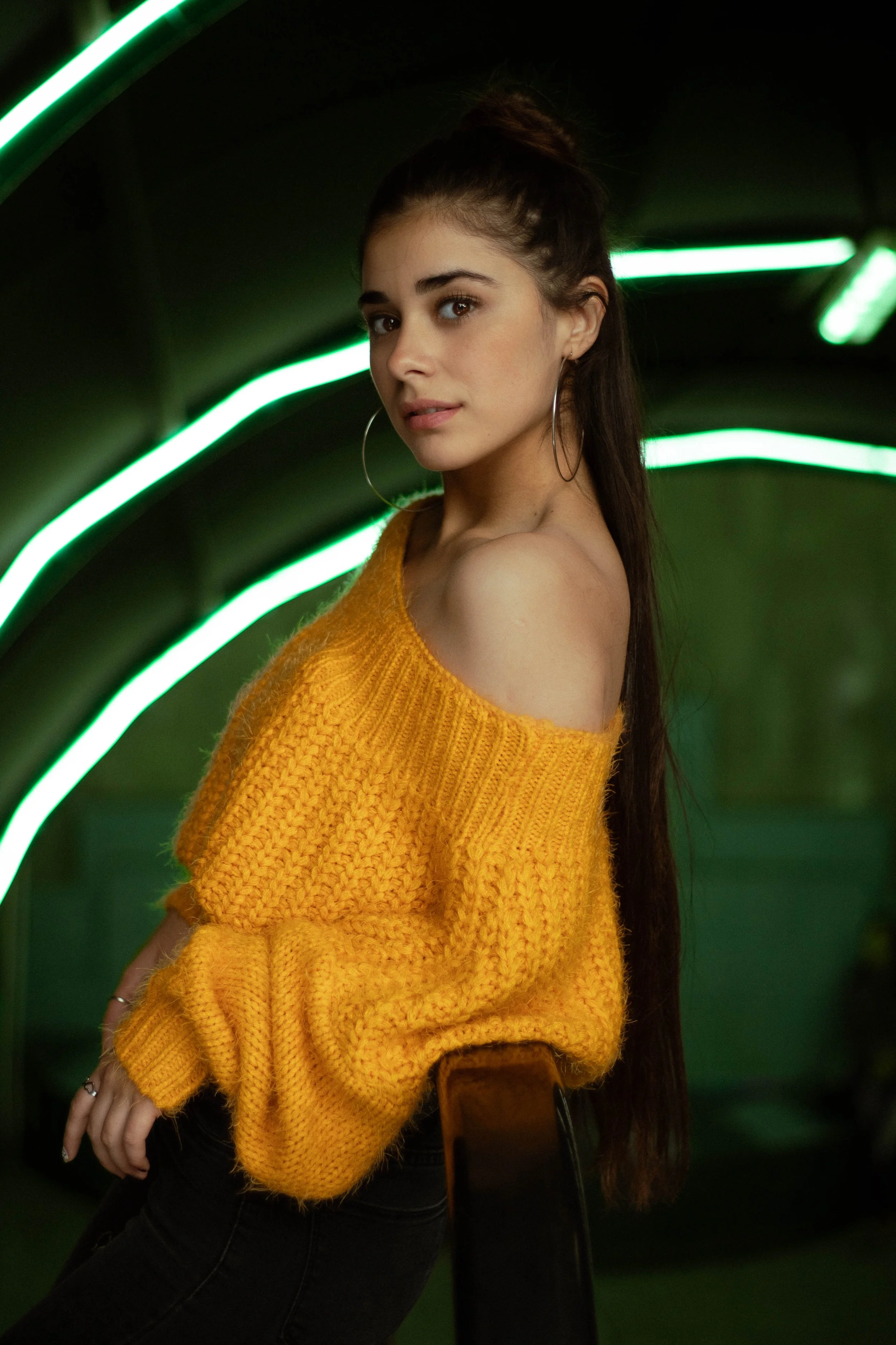 A young woman with long dark hair, wearing a yellow knitted off-shoulder sweater and large hoop earrings, sitting on a chair with a dark green background illuminated by neon green lights.
