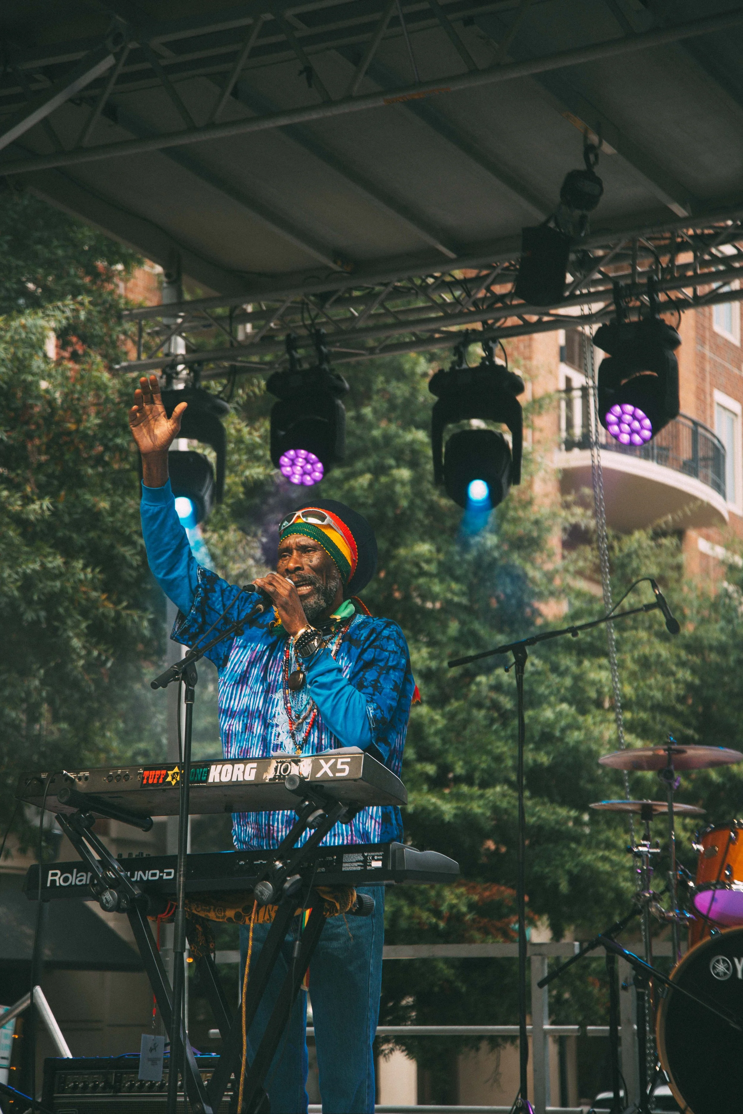 A male musician with a beard and dreadlocks, wearing a colorful headband and glasses, performs on an outdoor stage. He is singing into a microphone and playing a keyboard with various electronic gear. Stage lights illuminate him, and trees and buildi