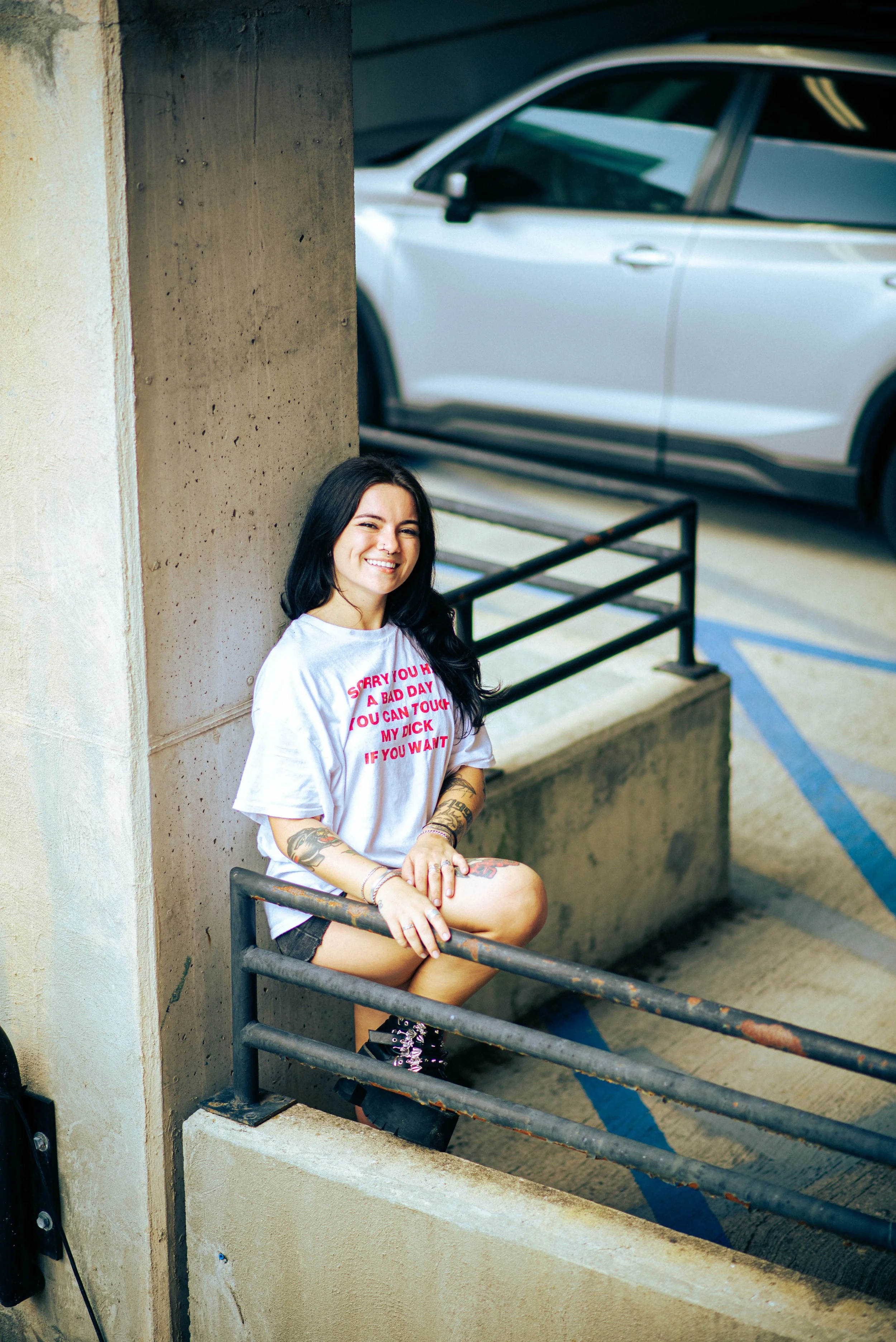A young woman with long dark hair, tattoos, and a nose piercing, sitting on a metal railing with one leg crossed over the other, smiling with teeth showing, in a parking garage next to a beige concrete wall with a gray car parked behind her.