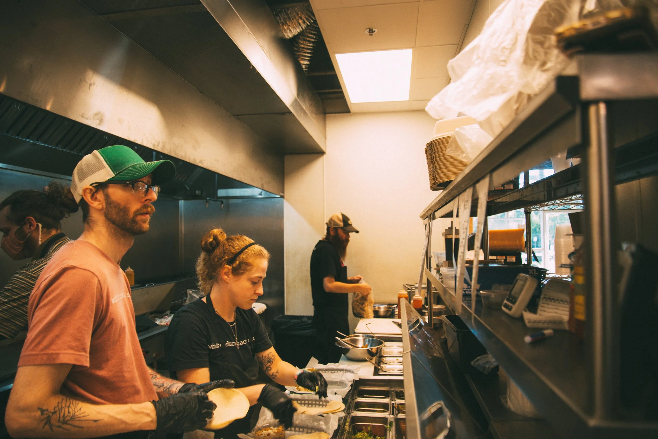 People working in a fast food restaurant kitchen preparing food, with shelves, containers, and kitchen equipment visible.