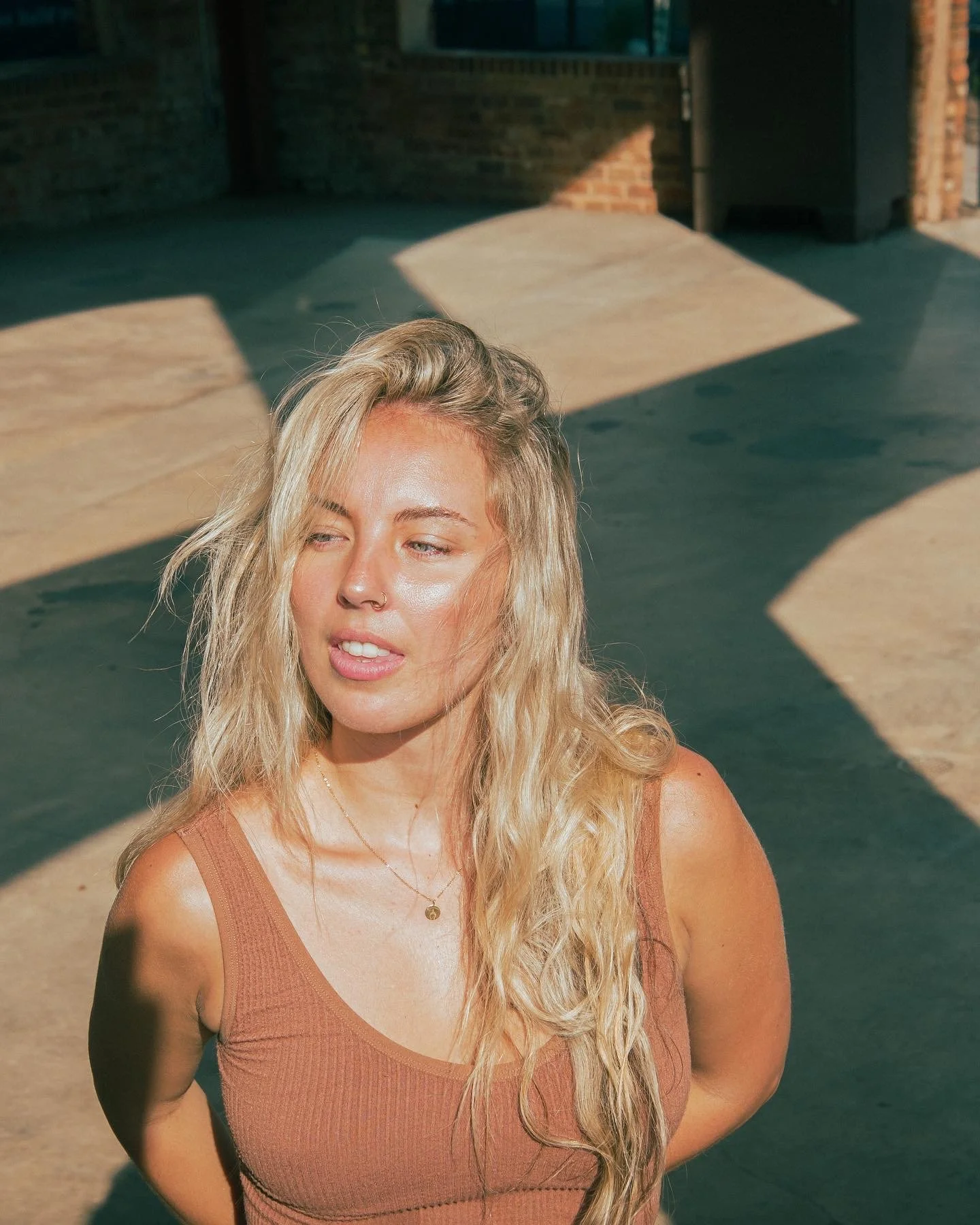 A young woman with long blonde hair, wearing a sleeveless top and a necklace, standing outdoors with sunlight casting shadows on the ground.