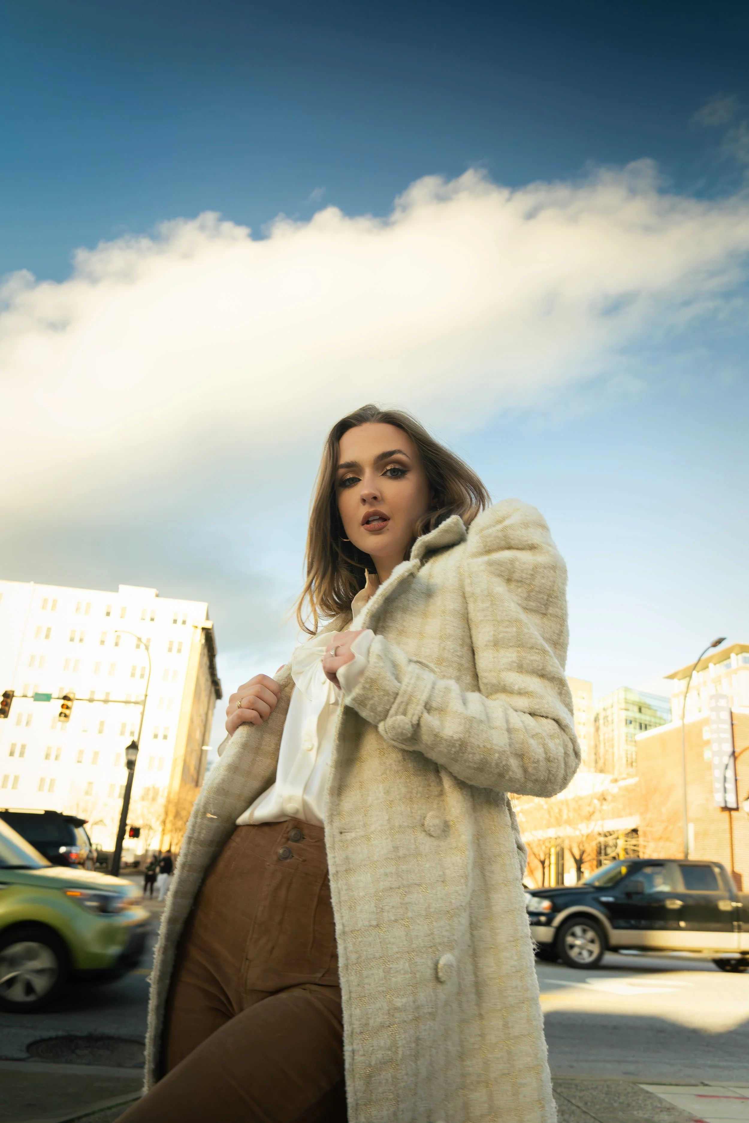 A woman with brown hair and makeup stands outdoors in a city, wearing a beige plaid coat over a white blouse and brown pants, with buildings and cars in the background under a partly cloudy sky.