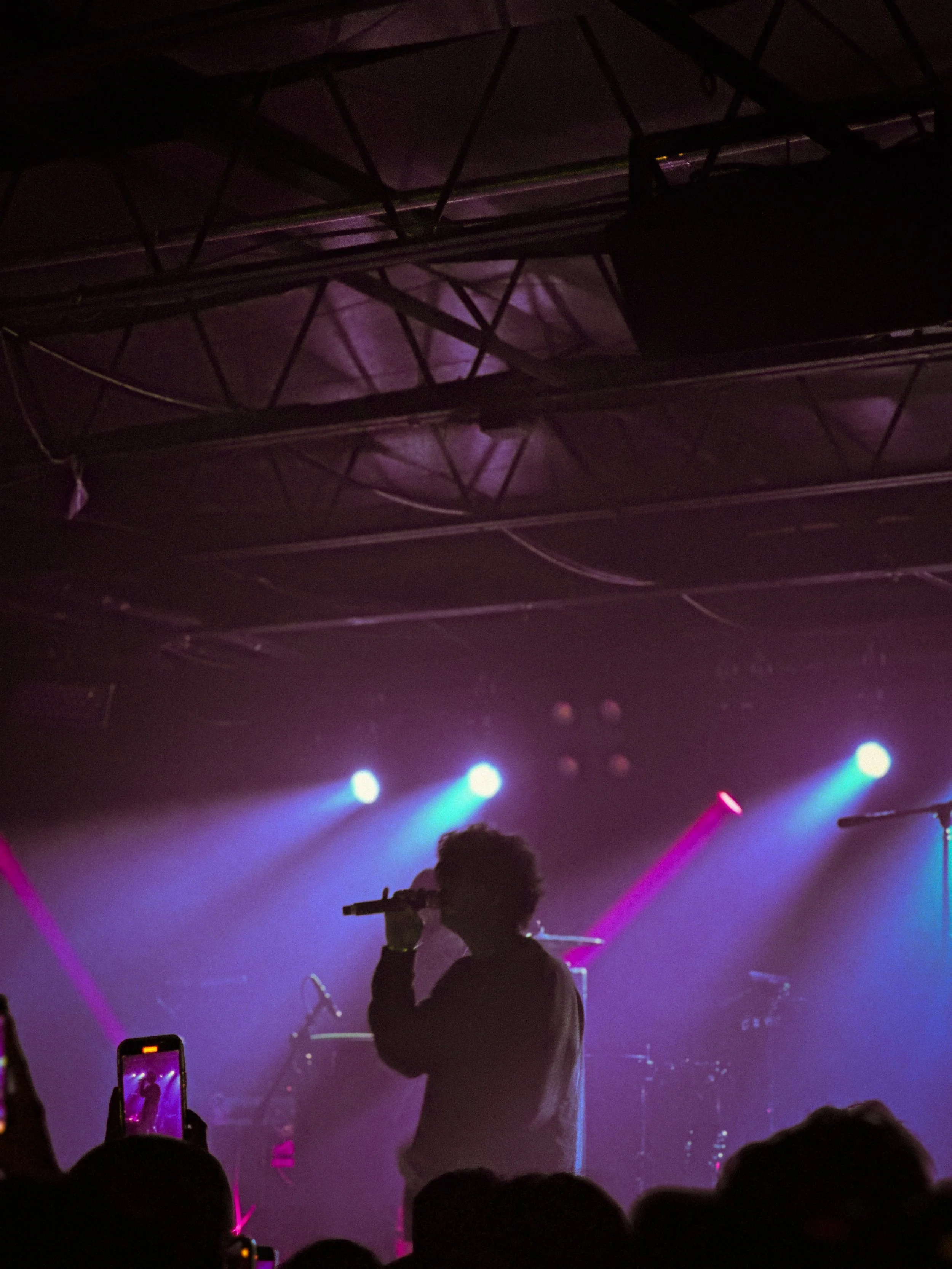 A performer on stage at a concert or club with colorful purple and blue lights, a drum set, and a person in front holding up a phone to record or take photo.