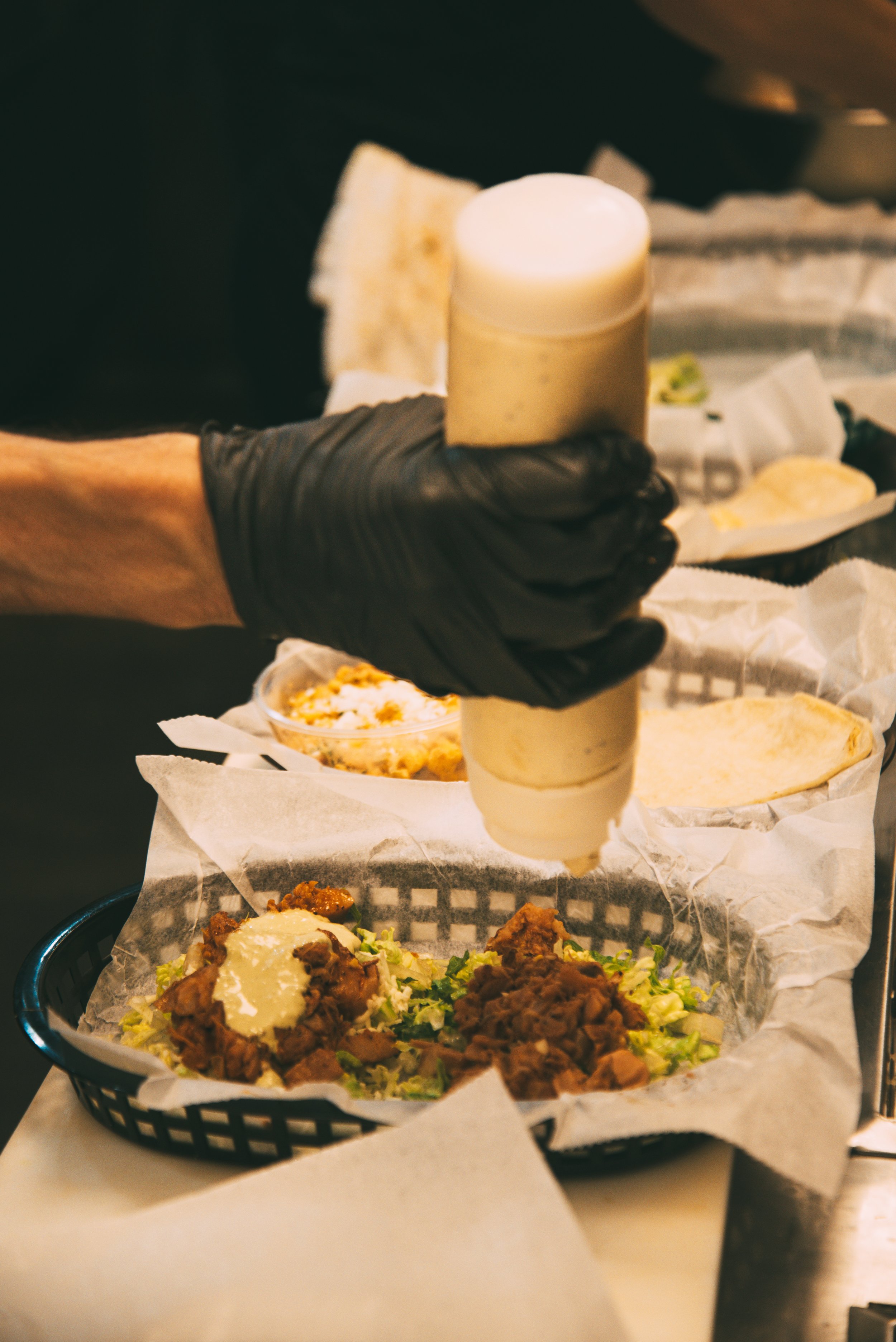 A person wearing black gloves holding a squeeze bottle over a plate of food. The plate contains shredded lettuce, cooked ground meat, grated cheese, and sour cream. In the background, there are various bowls and food items on a paper-lined tray or co