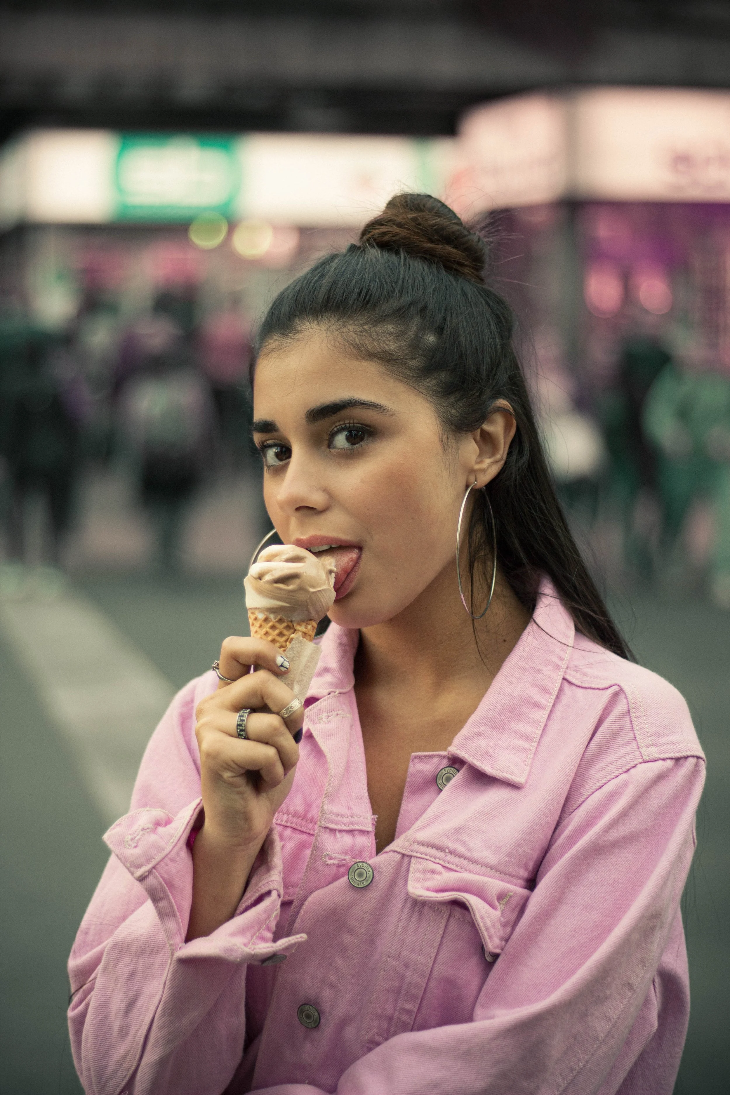 A young woman with dark hair in a high bun, wearing large hoop earrings and a pink jacket, is licking an ice cream cone while looking directly at the camera. The background appears to be a blurry indoor setting with bright lights.