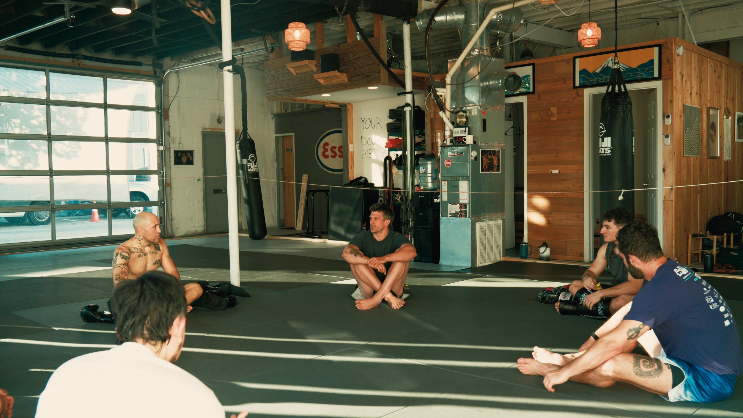 Group of men sitting on the floor in a martial arts or fitness studio with punching bags and wooden walls, engaged in a discussion or meditation.