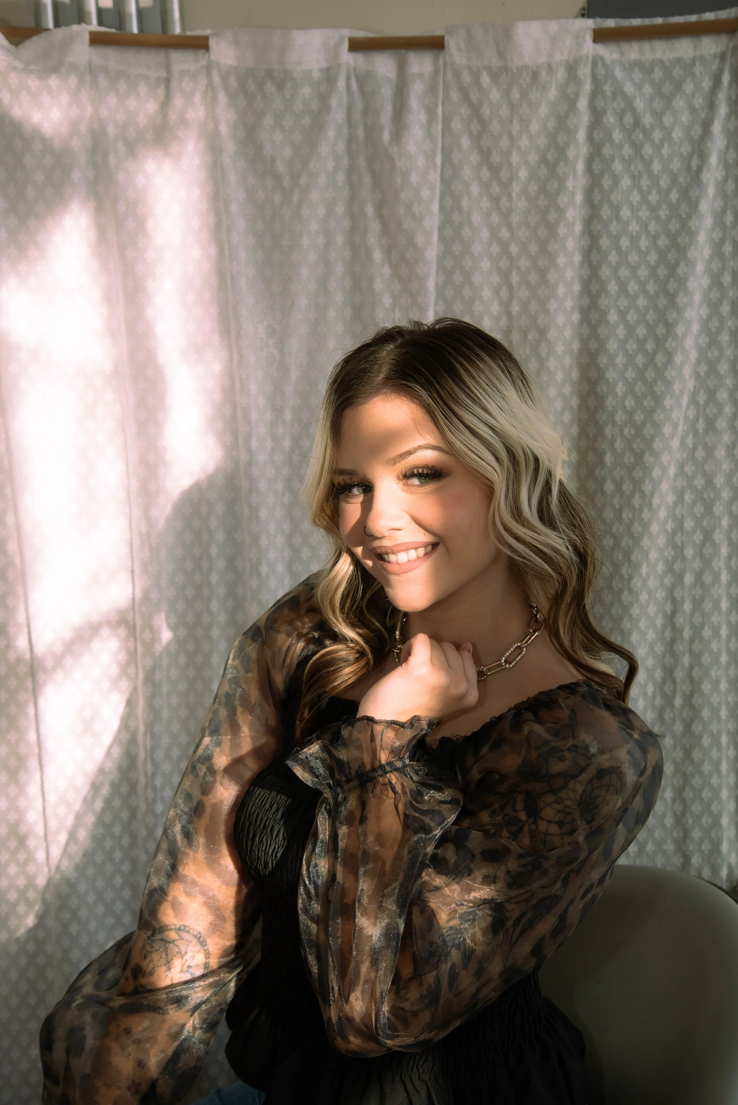 A woman with wavy blonde hair and tattoos on her arm, smiling and looking at the camera, standing in front of a light-colored patterned curtain, wearing a dark dress and a chain necklace.