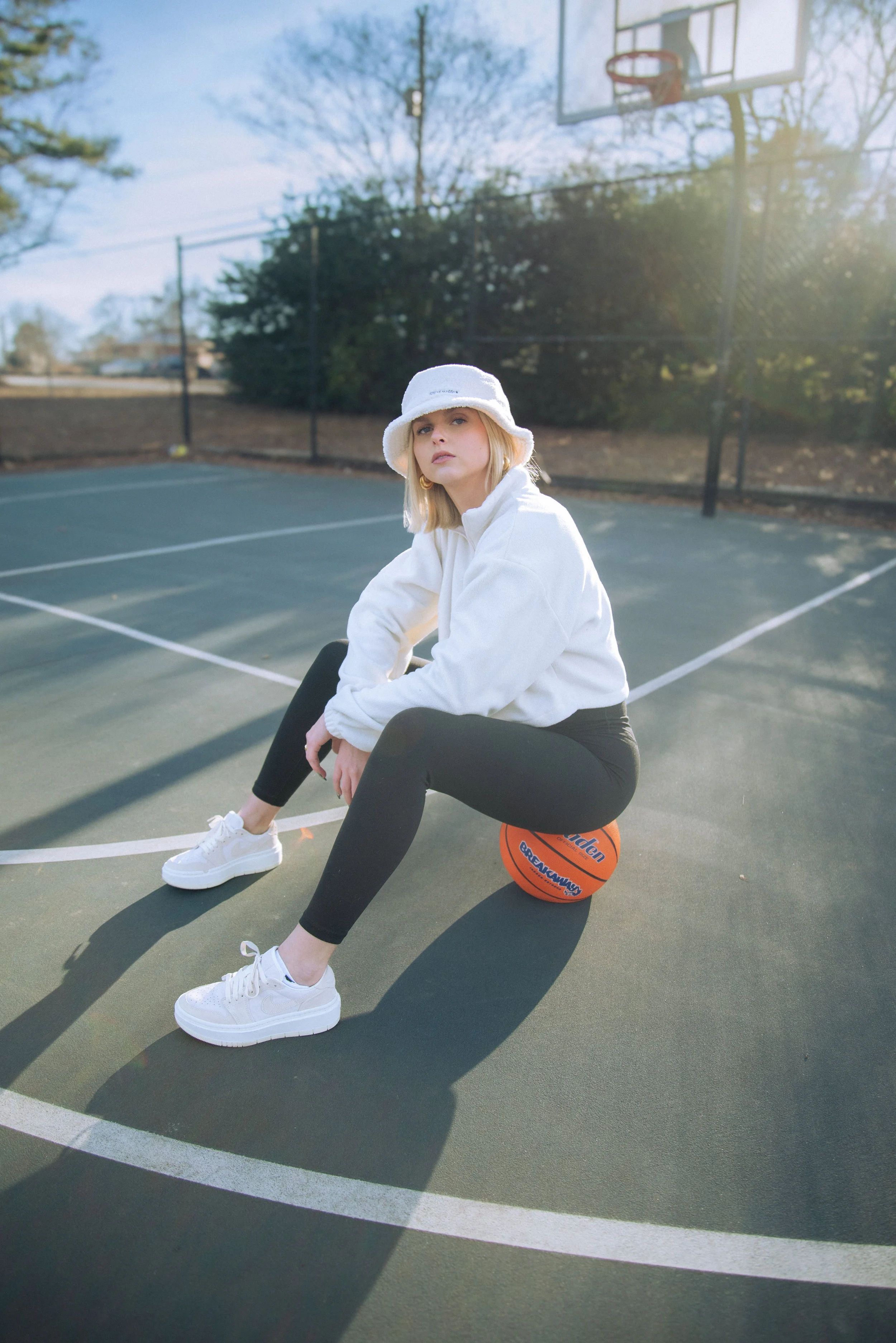 A young woman sitting on a basketball on an outdoor basketball court during daylight. She is wearing a white bucket hat, white jacket, black leggings, and white sneakers. The court is surrounded by trees and a fence, with a basketball hoop visible in