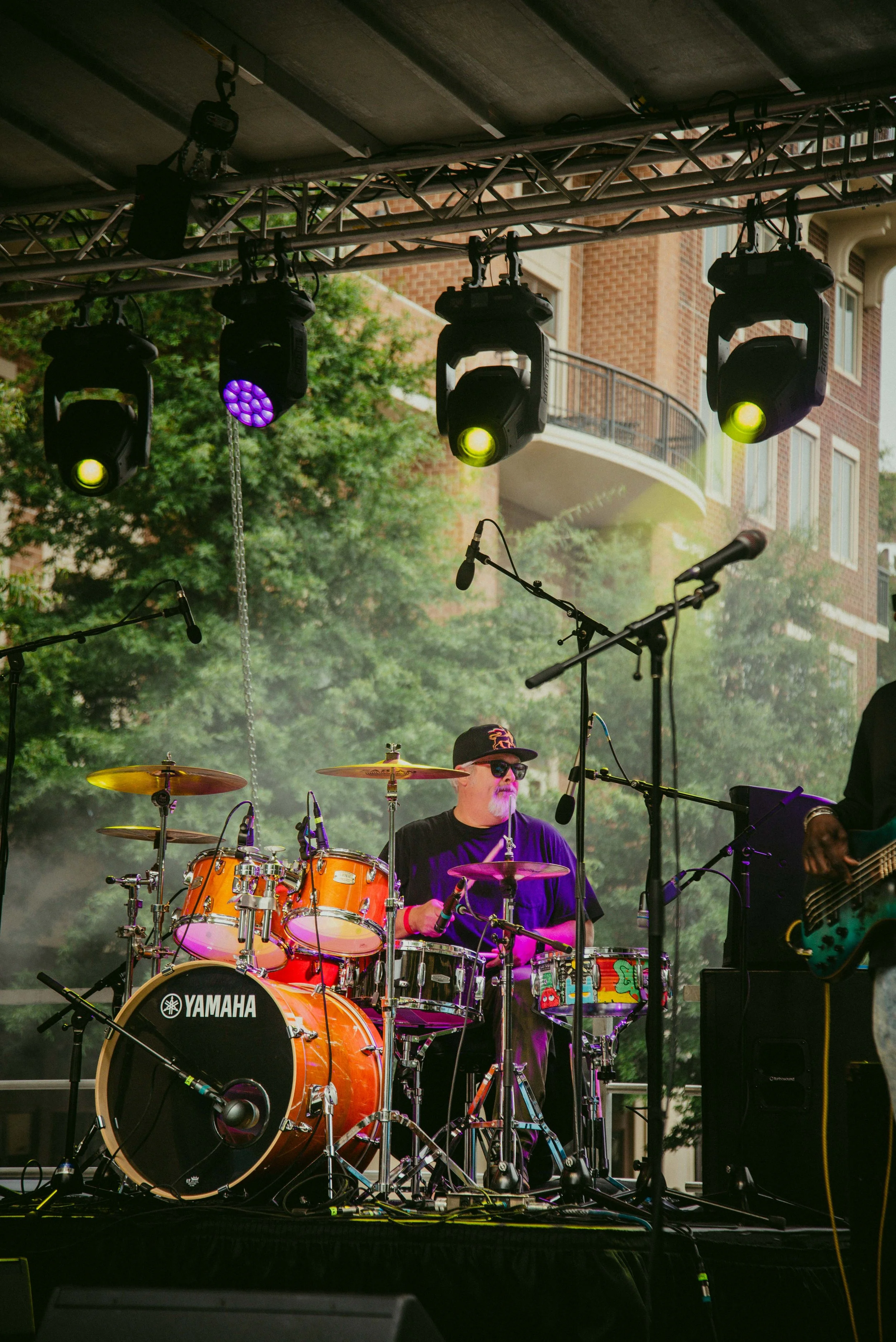 A live outdoor music performance featuring a drummer playing a Yamaha drum set and a guitarist in the background, under stage lights with trees and a building in the background.