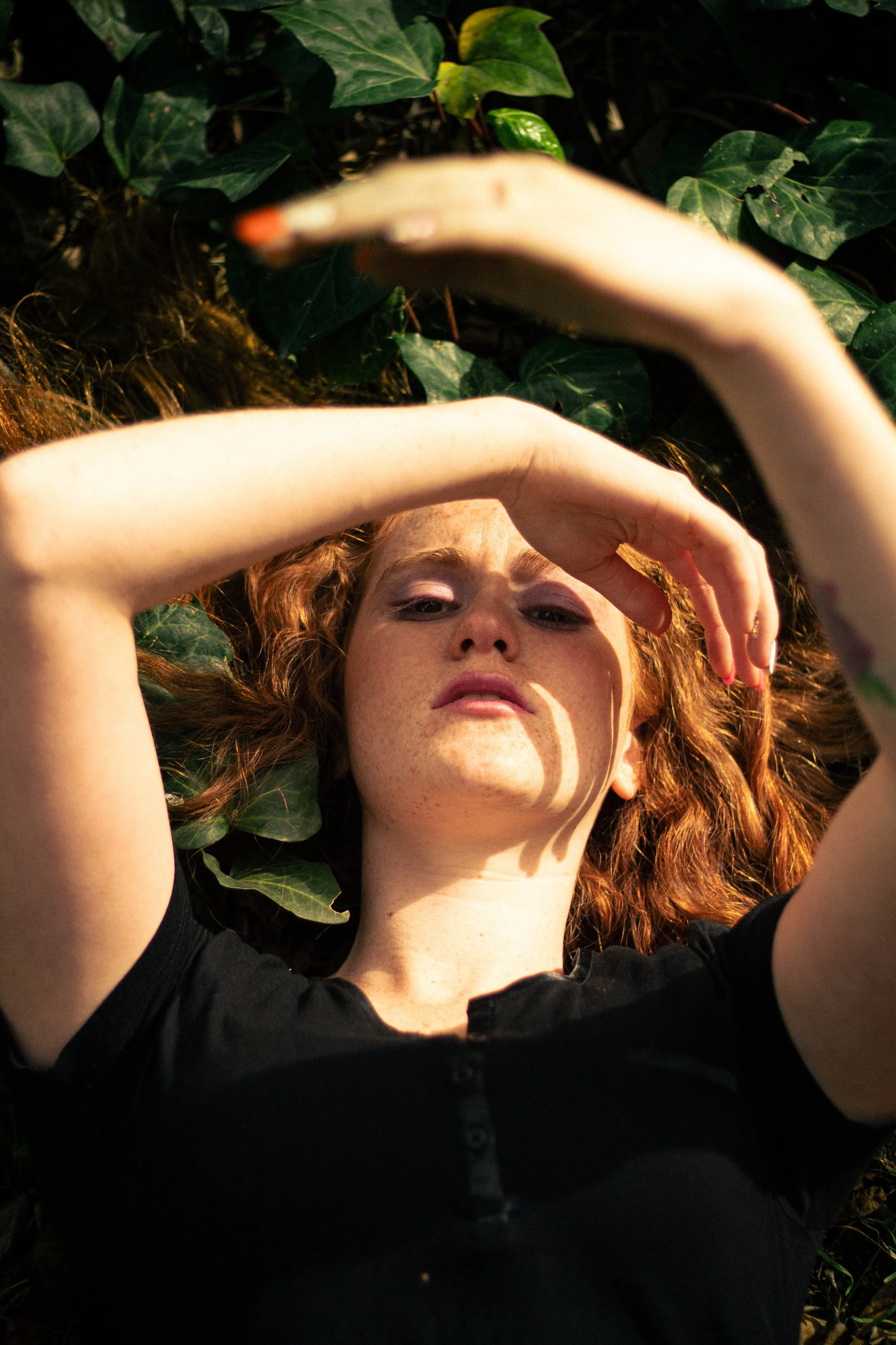 A woman with curly red hair lying on the ground surrounded by green ivy leaves, with her arm blocking part of her face.