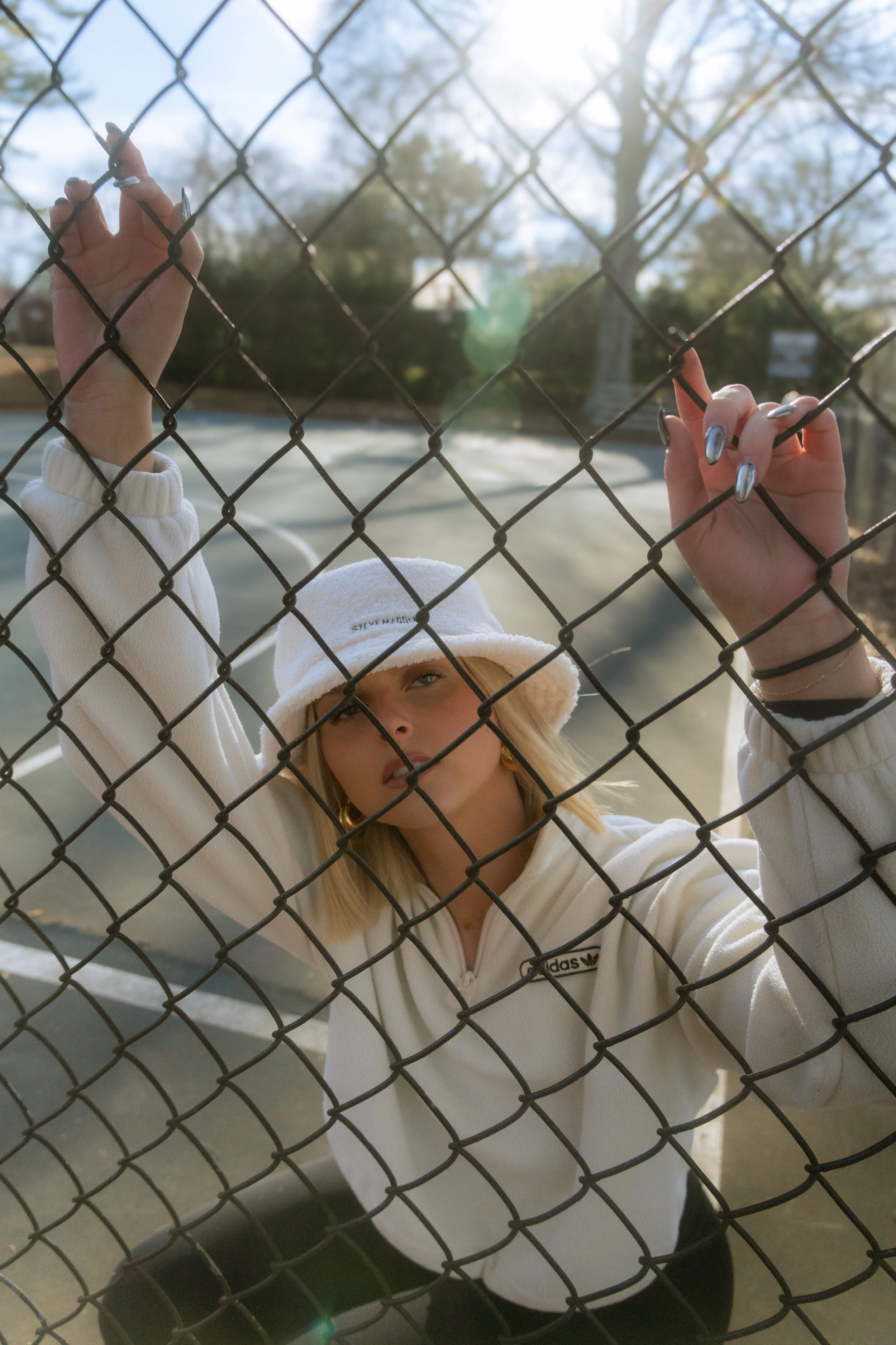A person with blonde hair wearing a white bucket hat and a white Adidas sweatshirt is holding onto a chain-link fence with both hands. The person has metallic silver painted nails and is standing outdoors, with trees and a tennis court surface in the