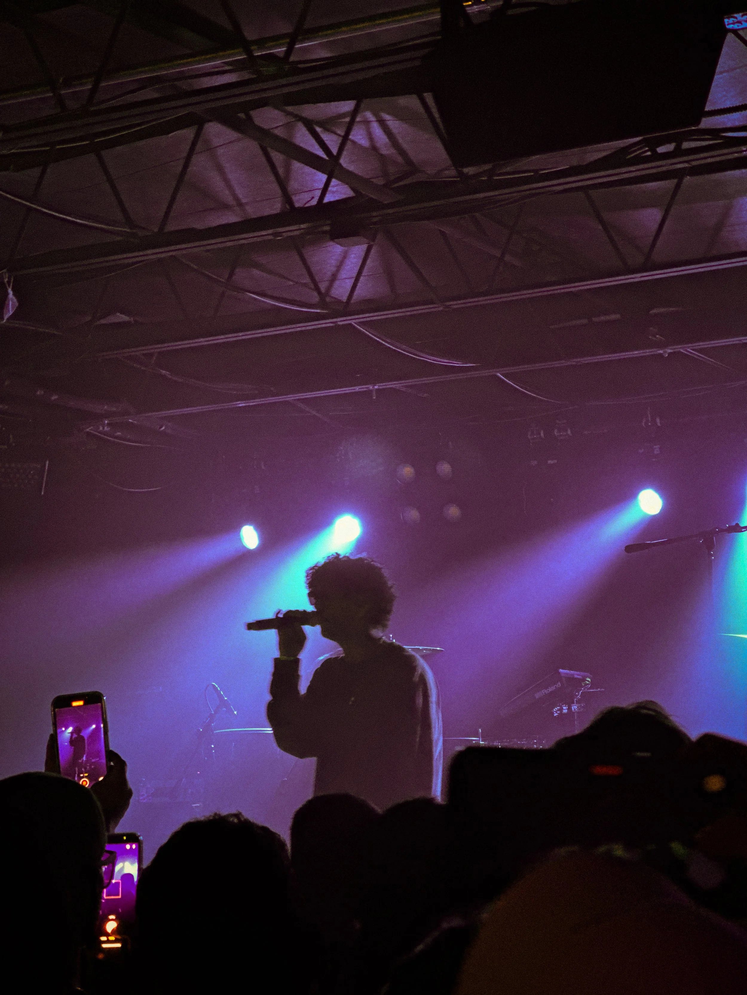 A silhouette of a person singing into a microphone on stage, with stage lights and audience members capturing the scene with phones, in a concert or live performance setting.