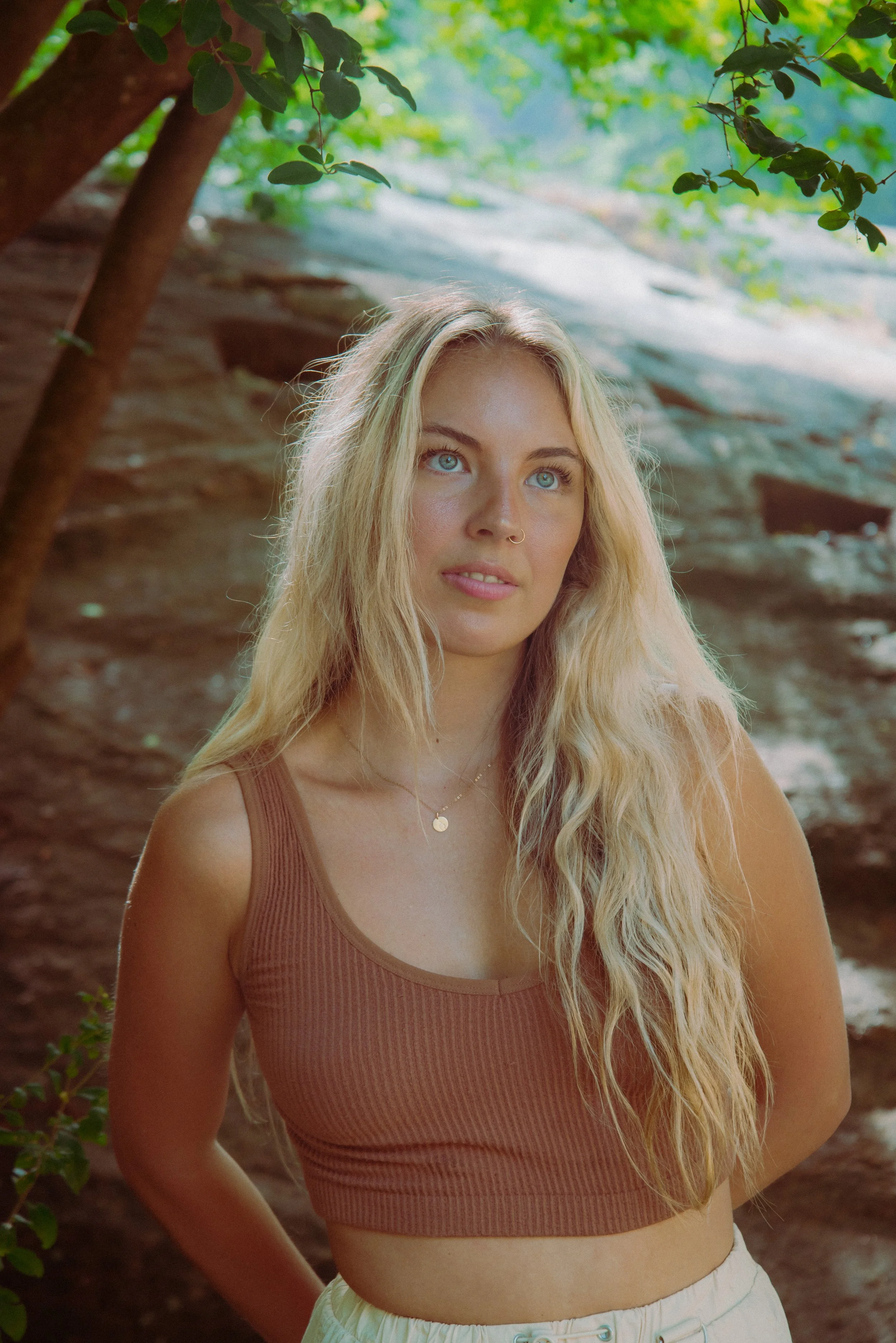 A young woman with long blonde hair, blue eyes, and a nose piercing looking slightly upward. She is wearing a ribbed brown crop top and white shorts, standing outdoors near a tree with green foliage and a rocky background.