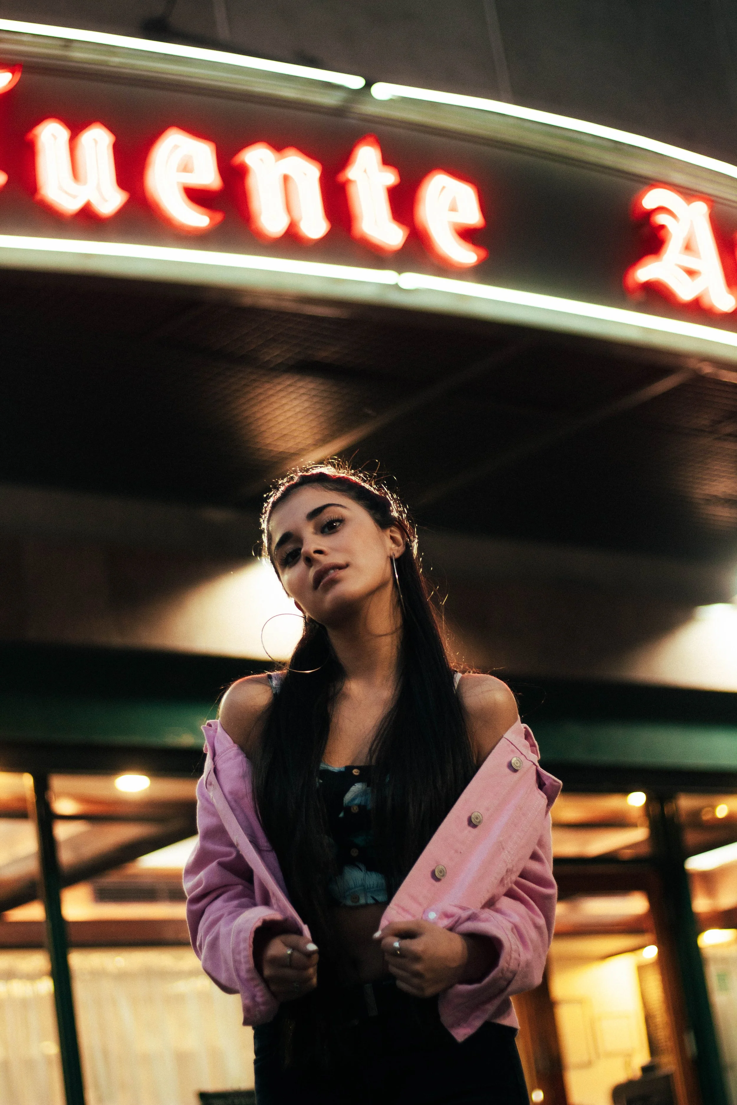A young woman with long dark hair wearing a pink jacket, standing in a dimly lit indoor space, with a neon sign that says 'Lente' in the background.