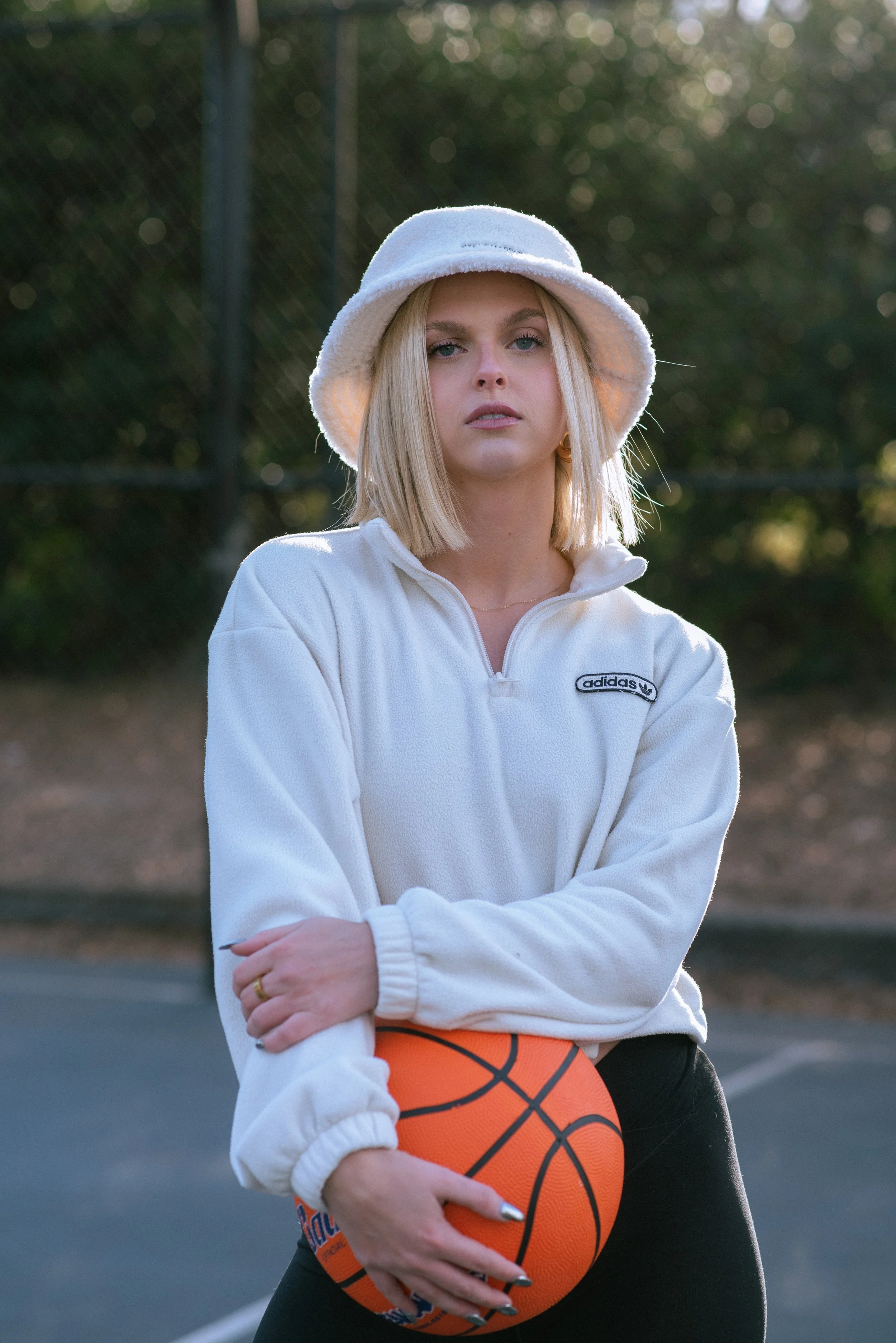 A woman standing outdoors holding an orange basketball, wearing a white hat and a white zip-up jacket, with a blurred background of a basketball court and trees.