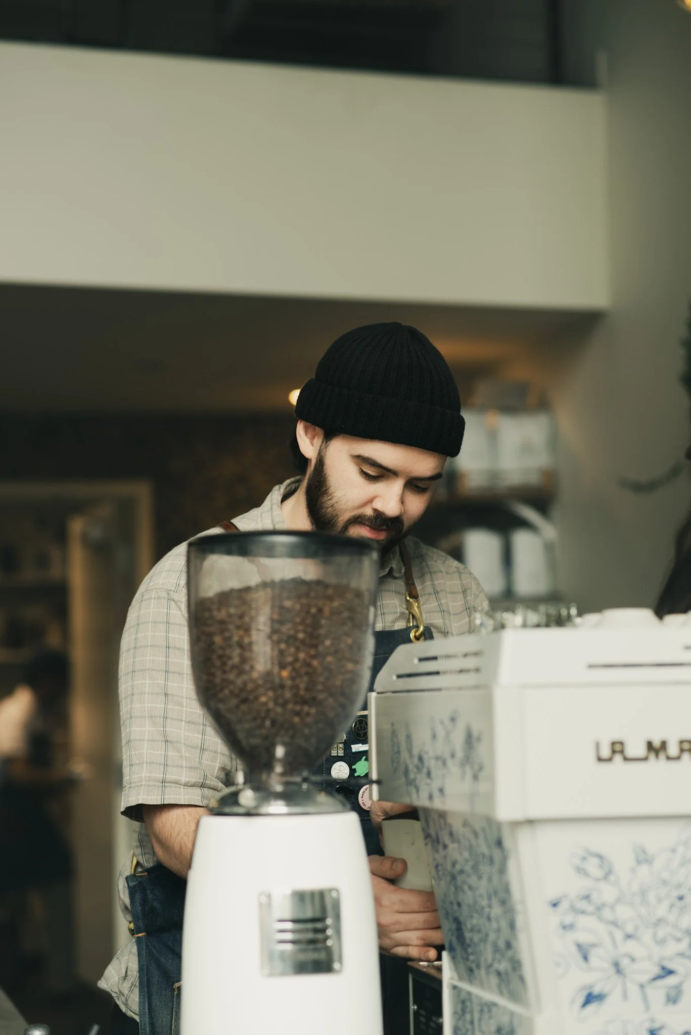 A barista with a beard and a beanie preparing coffee behind a counter, with coffee equipment and containers in the foreground.