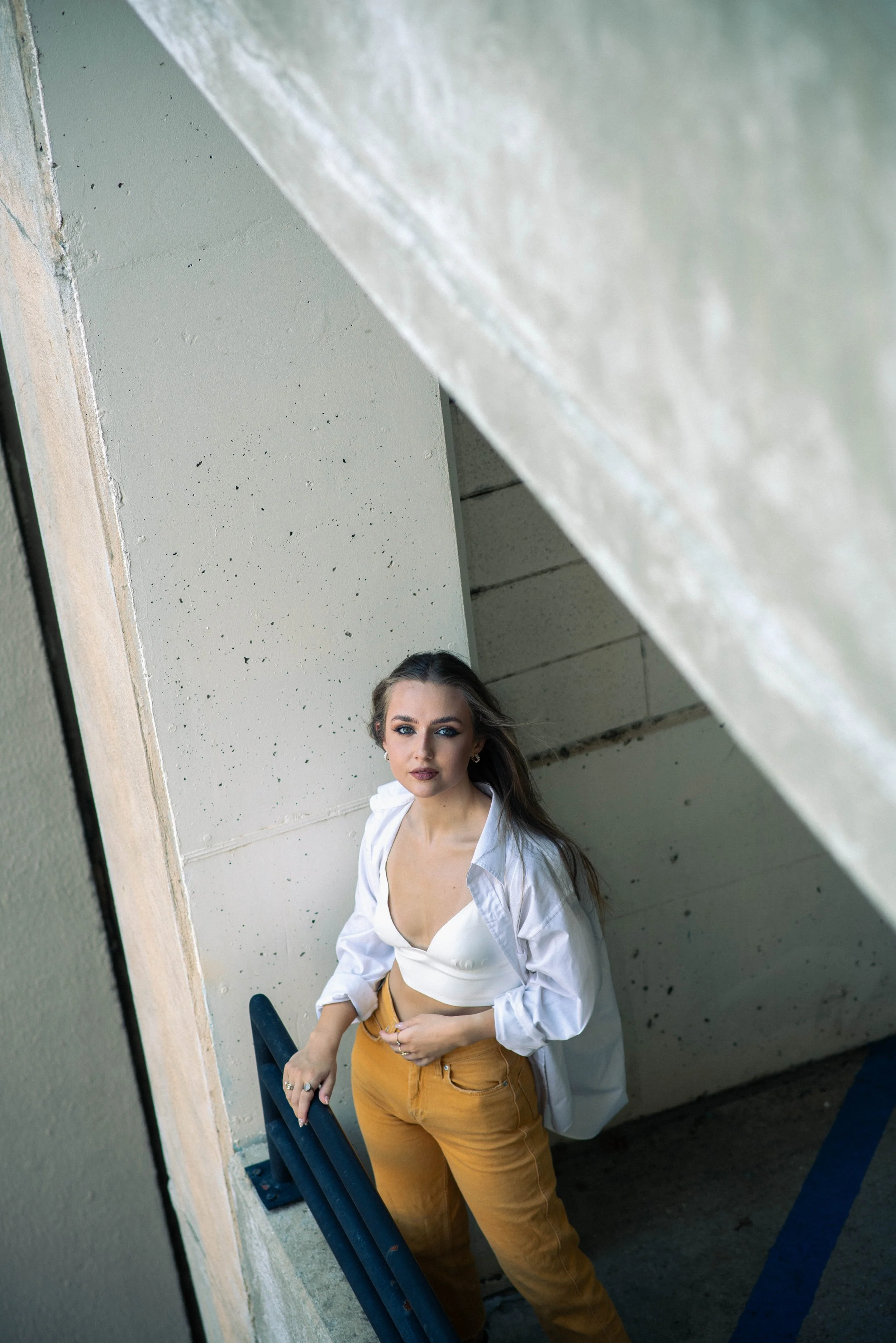 A young woman stands on a staircase outside, wearing a white blouse and mustard-colored pants, looking directly at the camera. The photo is taken through a reflective surface, creating a slightly distorted perspective.