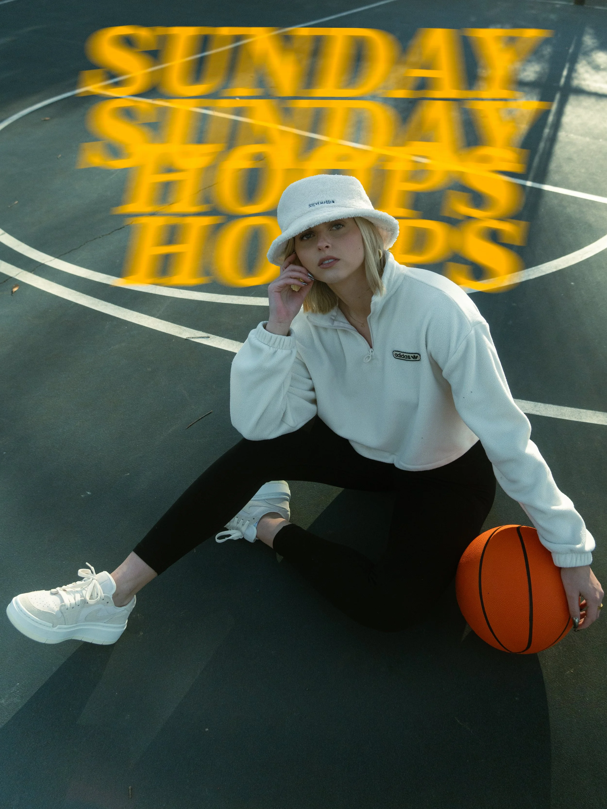 A young woman sitting on a basketball court with an orange basketball, wearing a white bucket hat, white Adidas sweatshirt, black pants, and white sneakers, with the word "SUNDAY" in yellow blurry text above her.