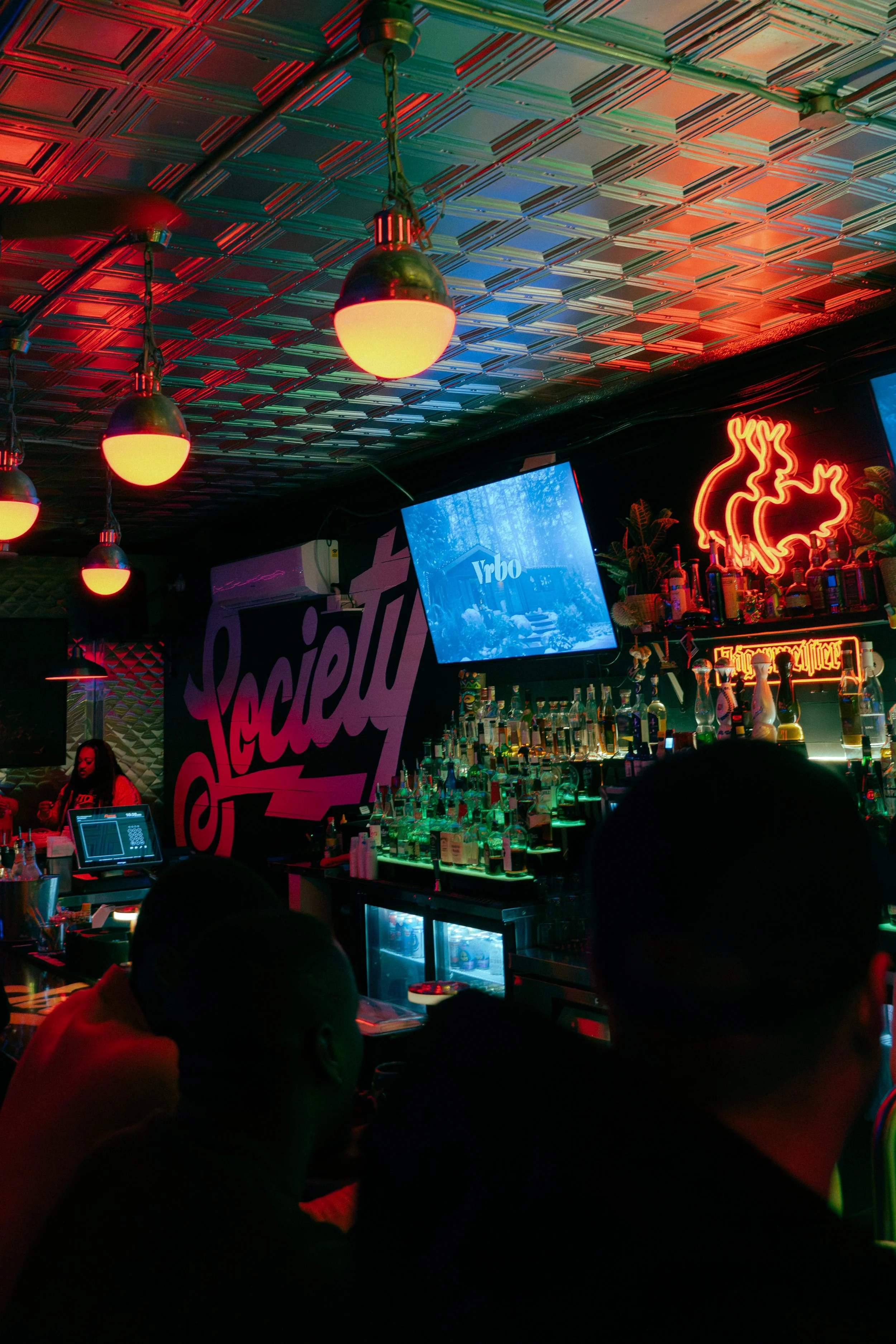The interior of a bar with neon lighting, including a neon pig and neon text, a television screen displaying a forest scene, and bottles of alcohol on shelves. The ceiling has a metallic, patterned surface with hanging lights.
