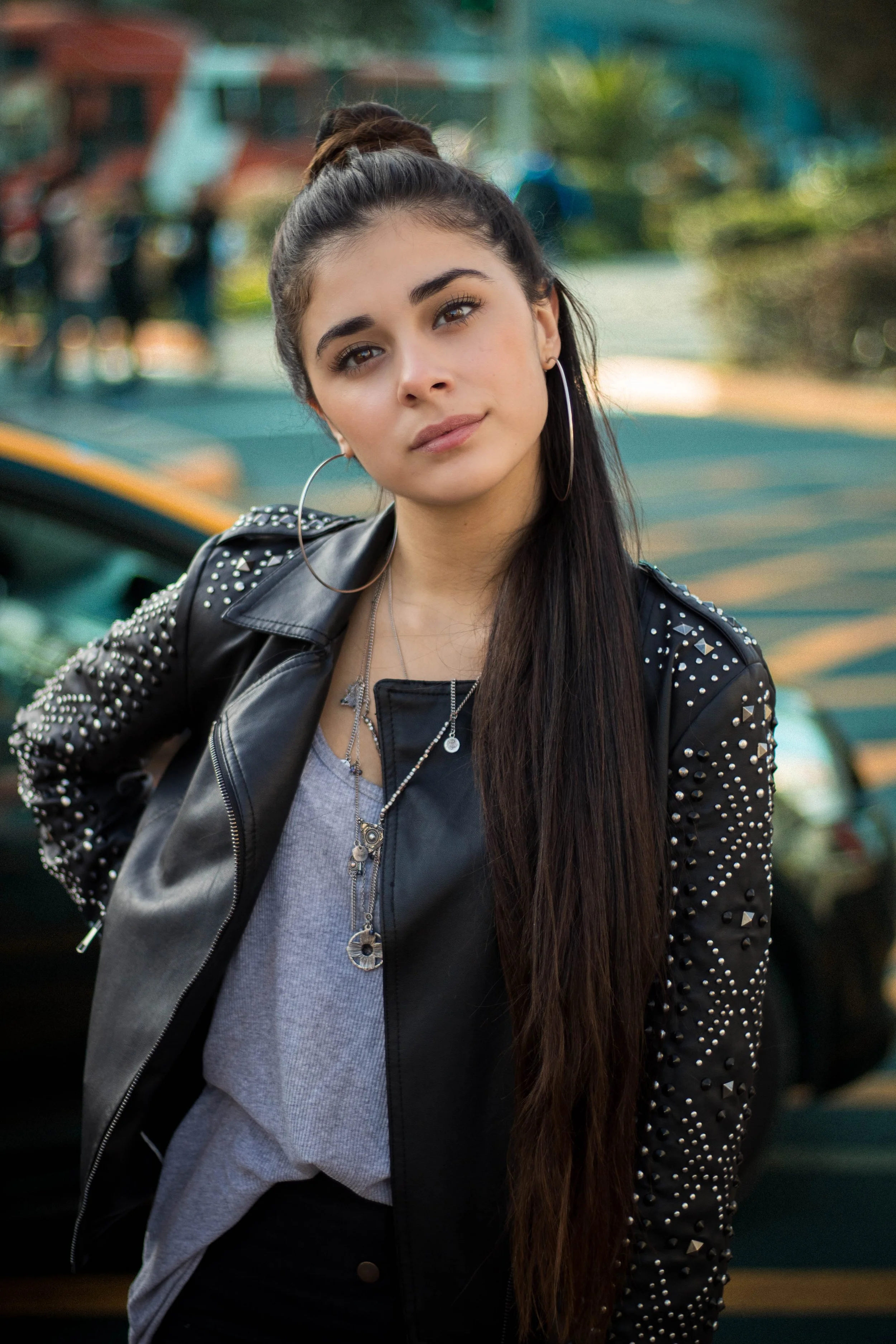 A young woman with long dark hair in a high bun, wearing hoop earrings, a leather jacket with studs, and layered necklaces, outdoors with a blurred background including trees and a vintage car.