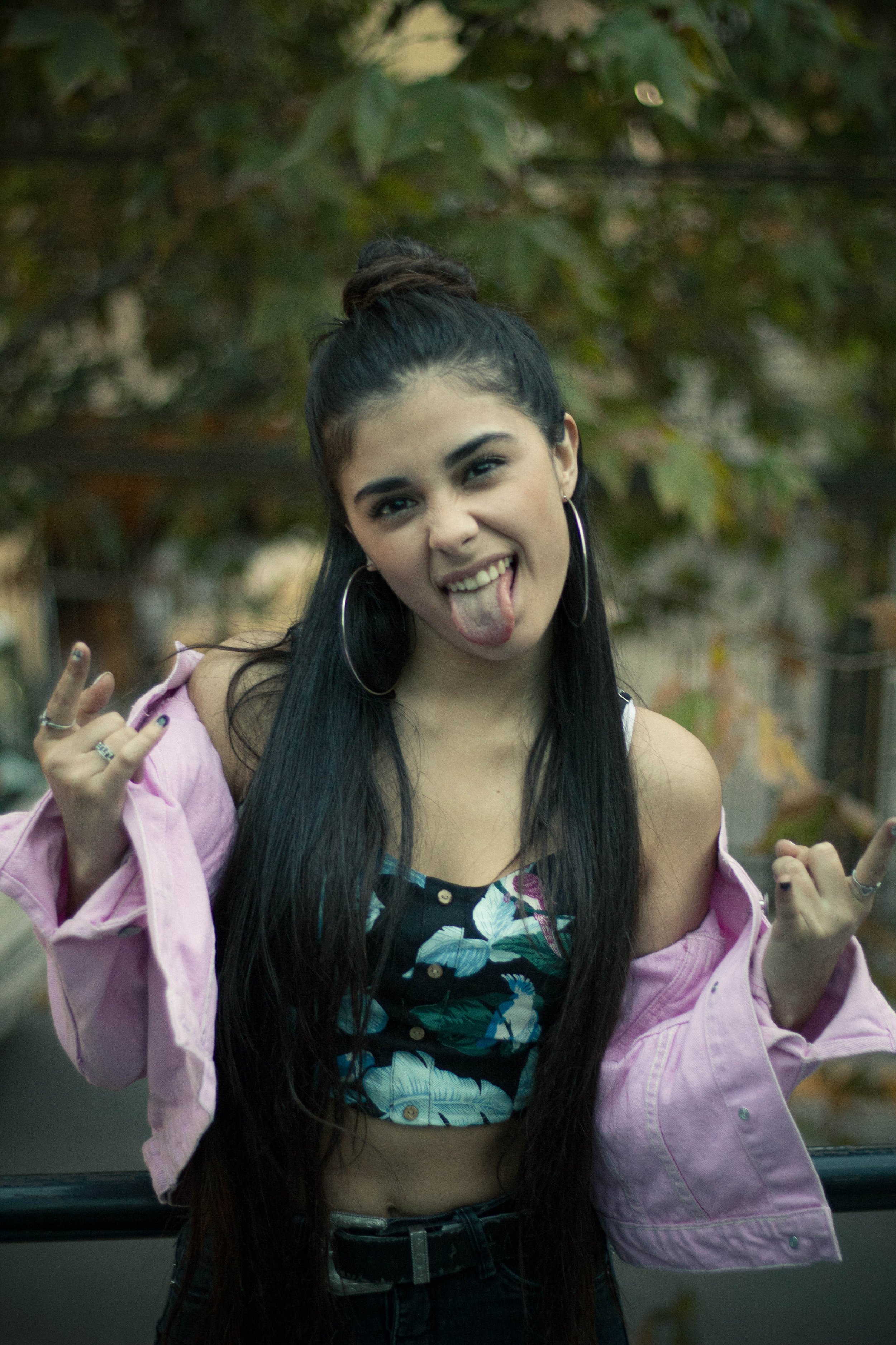 Young woman with long dark hair in a bun, sticking out her tongue, making a rock and roll hand gesture, wearing hoop earrings, a floral crop top, and a pink jacket, outdoors with green foliage in the background.