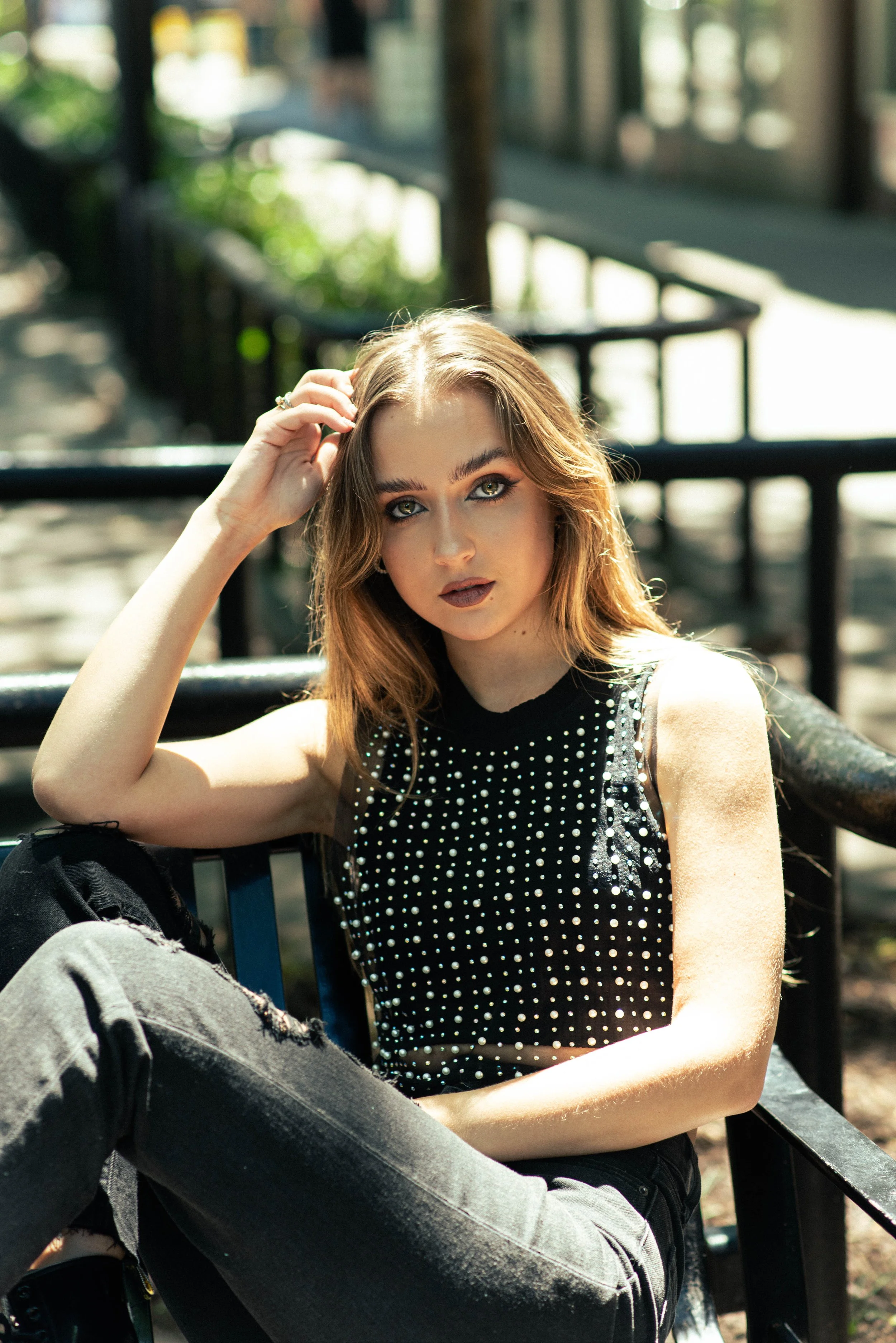 A young woman with long, light brown hair and gray eyes, wearing makeup, a black sleeveless top with shiny embellishments, and ripped black jeans, sitting on a park bench outdoors on a sunny day.