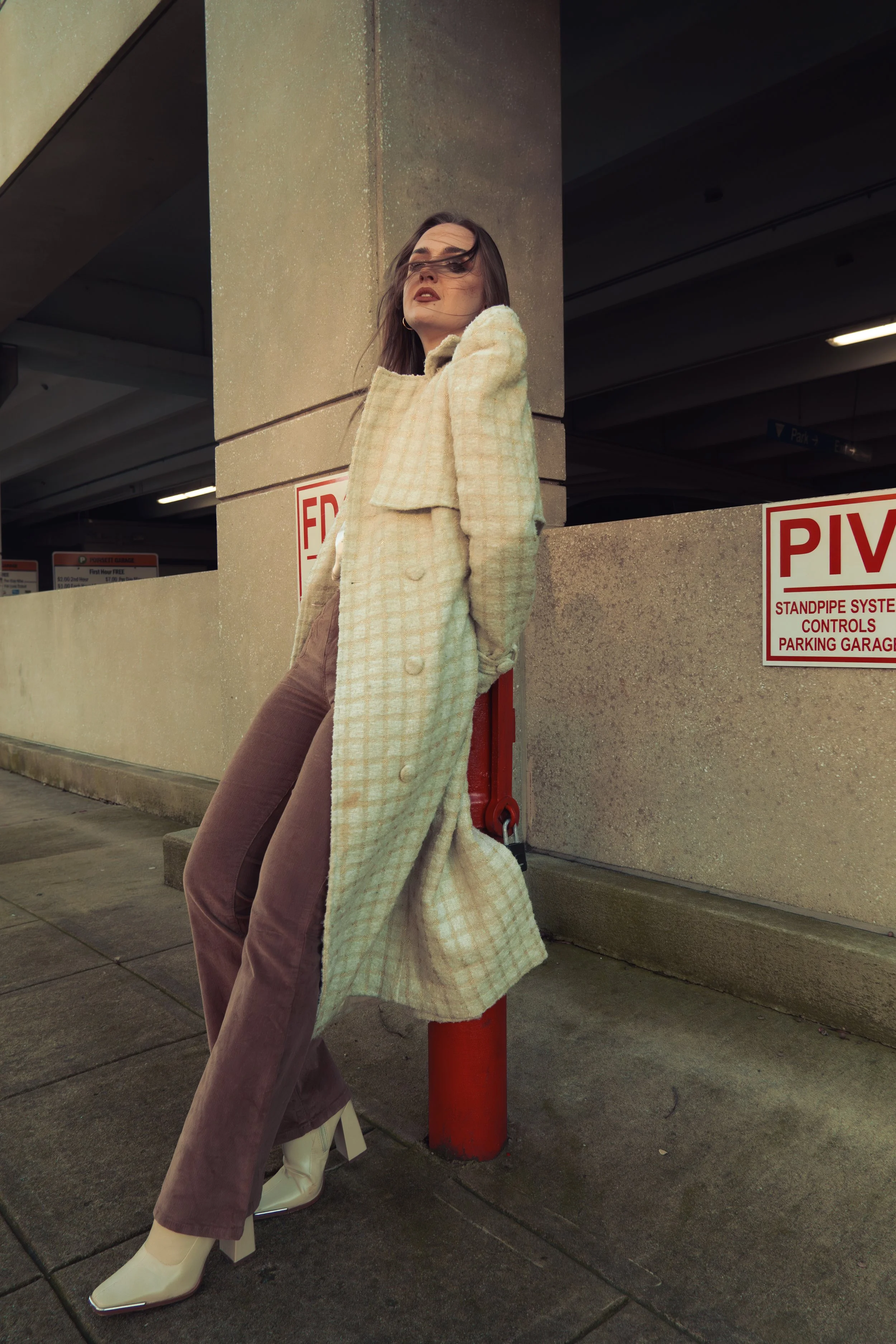 A woman leaning against a red post in a parking garage, wearing a beige checkered coat, brown pants, and cream boots.