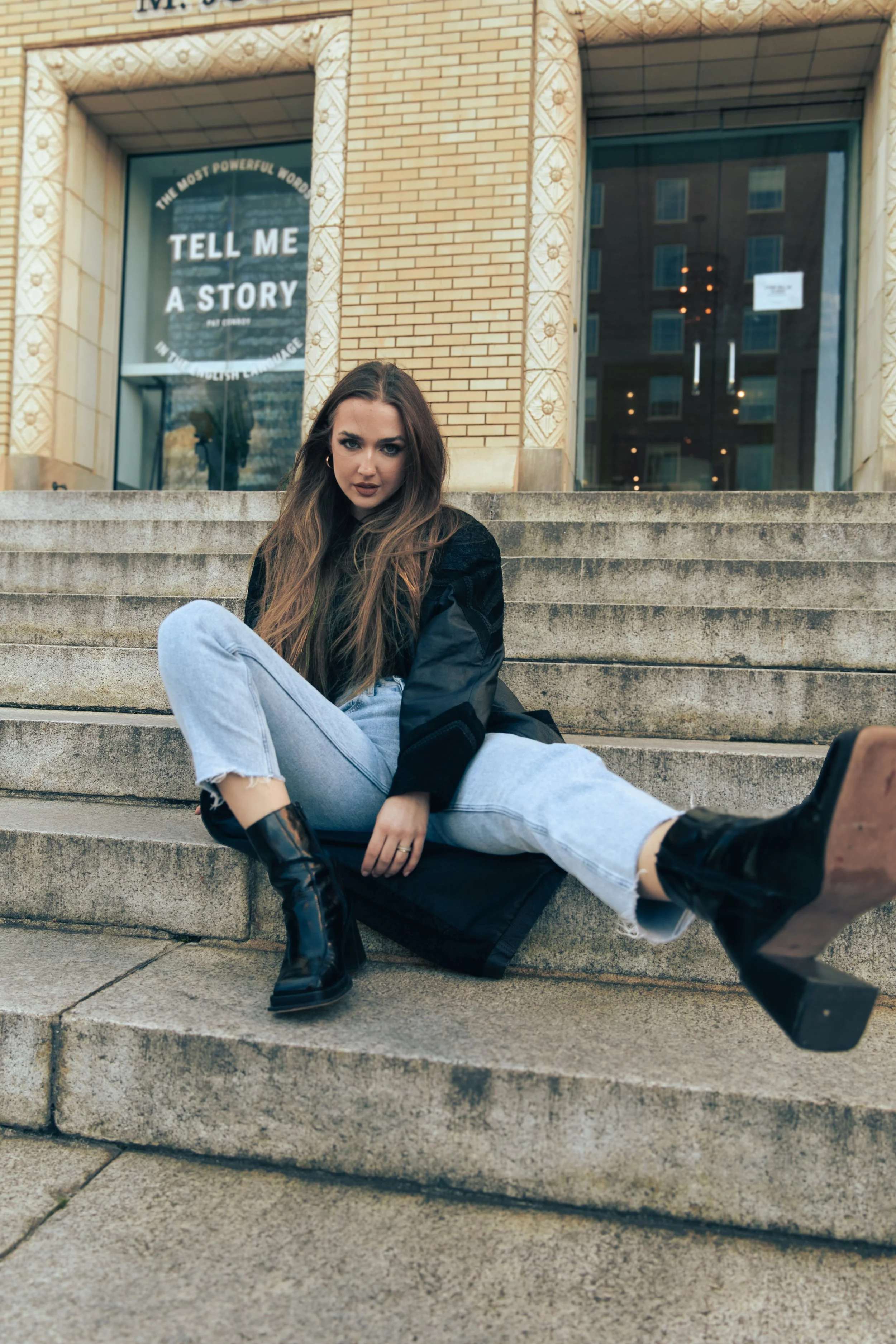 A woman with long brown hair sitting on outdoor steps, wearing a black jacket, light blue jeans, and black boots, in front of a building with a glass door and a window that reads 'Tell me a story'.