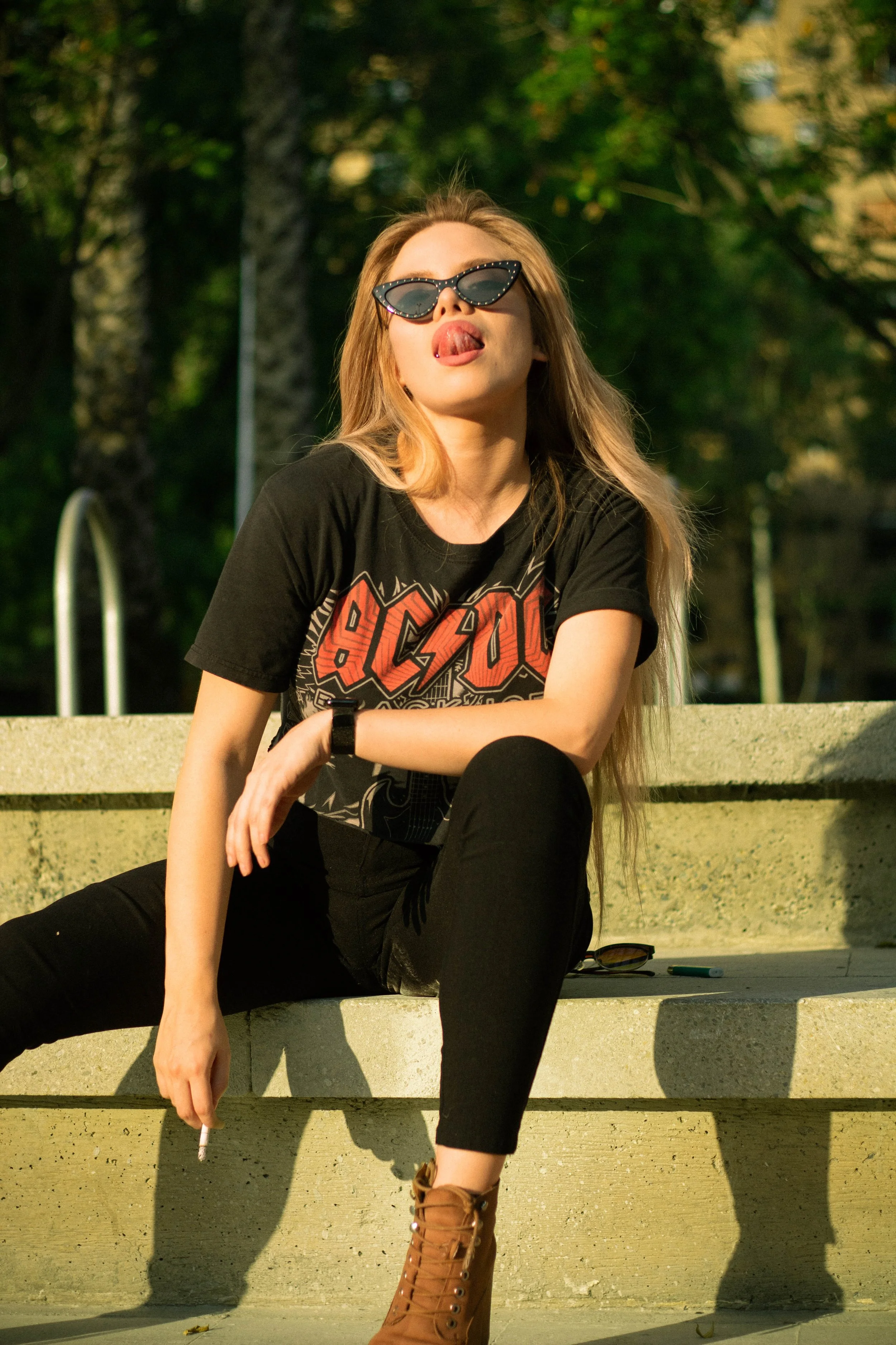 A young woman with long blonde hair wearing sunglasses, a black AC/DC t-shirt, black pants, and brown lace-up boots, sits on a concrete park bench in sunlight with trees in the background, sticking her tongue out.